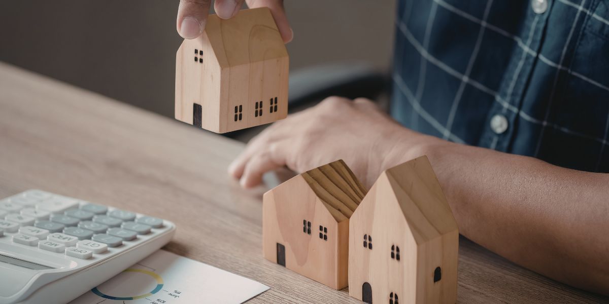 A person placing a small wooden house model on a desk next to two other house models, a calculator, and documents.
