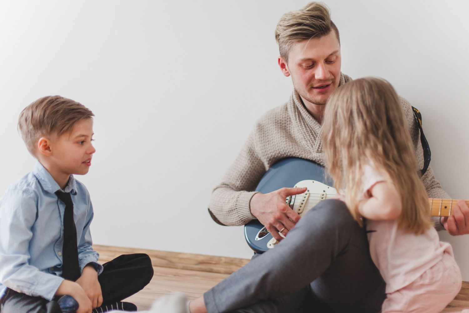 A man is playing a guitar to two children while sitting on the floor.