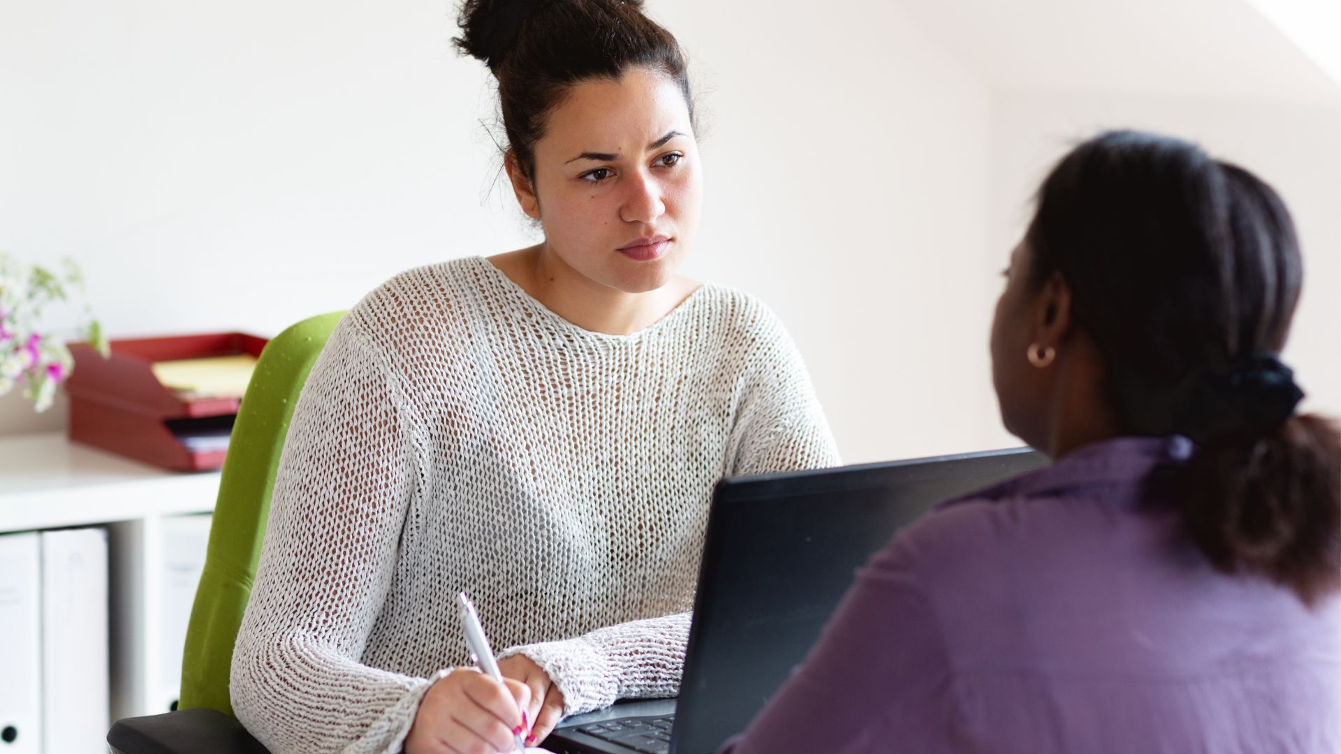 Two women are sitting at a desk with a laptop and talking to each other.