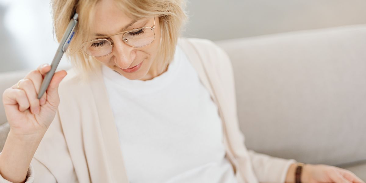 Woman with glasses and blonde hair holding a pen, looking down in thought. She is wearing a white shirt and beige cardigan, and seated on a couch.