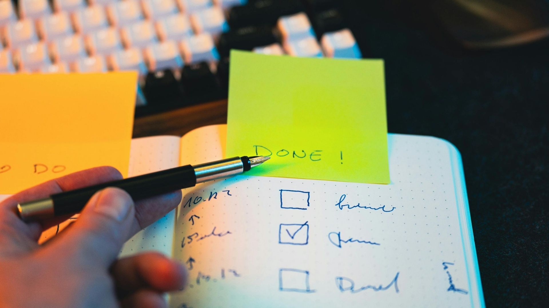 Hand writing a checklist in a notebook beside a yellow sticky note and computer keyboard