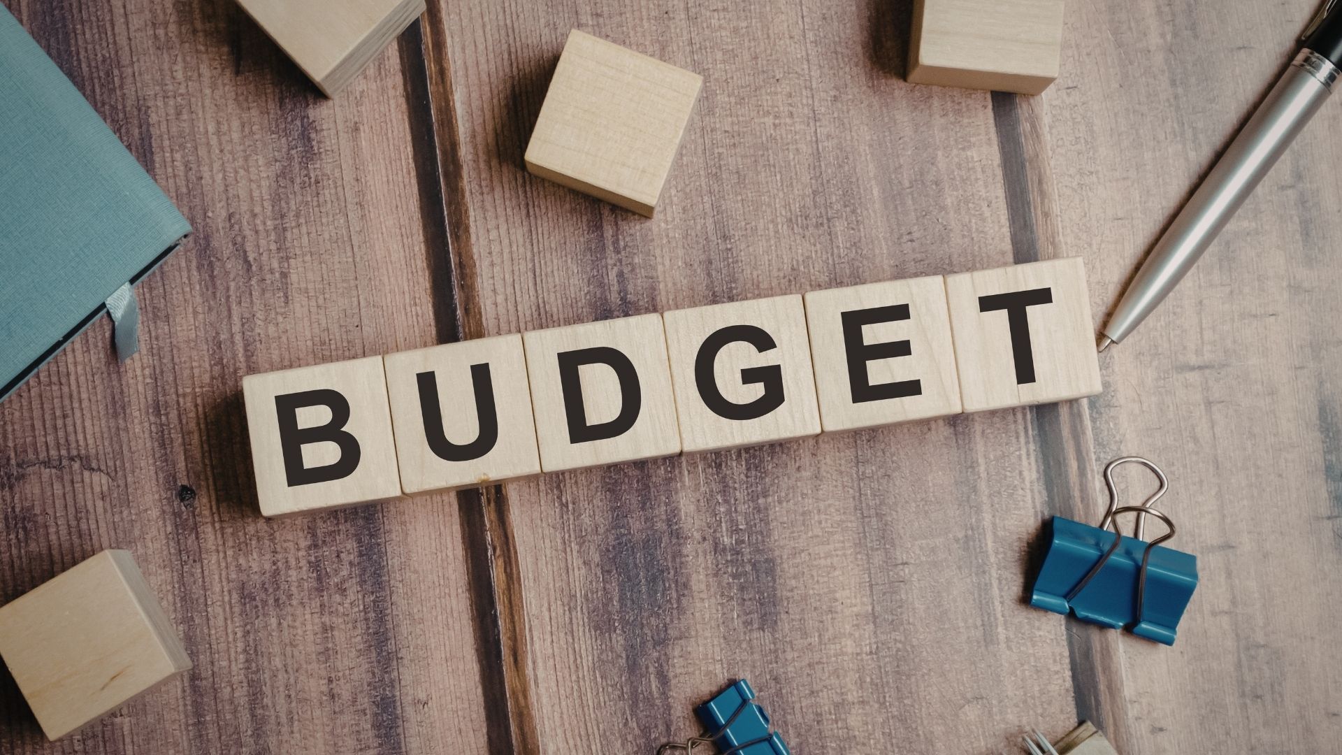 Scrabble tiles spelling “BUDGET” on a wooden desk with binder clips and sticky notes