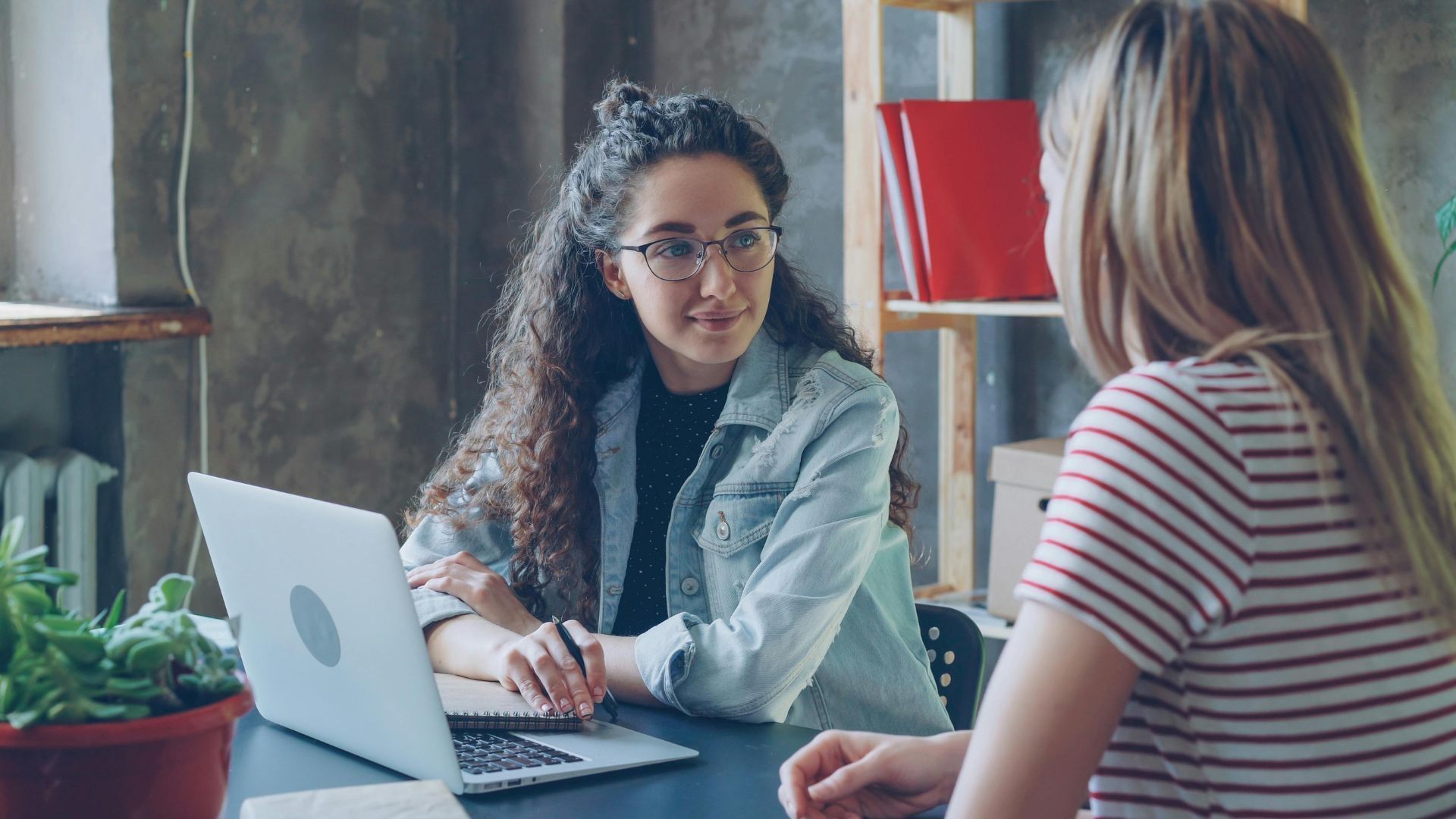 Two people discuss work at a table with a laptop in a modern office.
