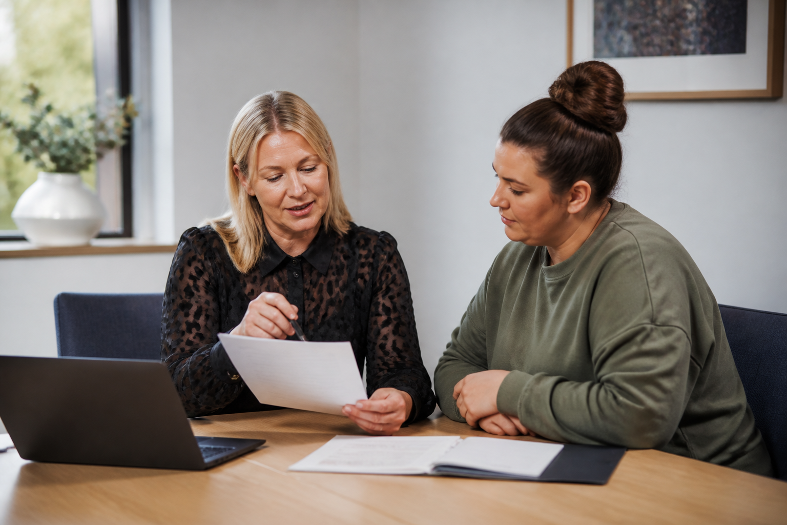 Two women review documents at a table with a laptop in a bright office
