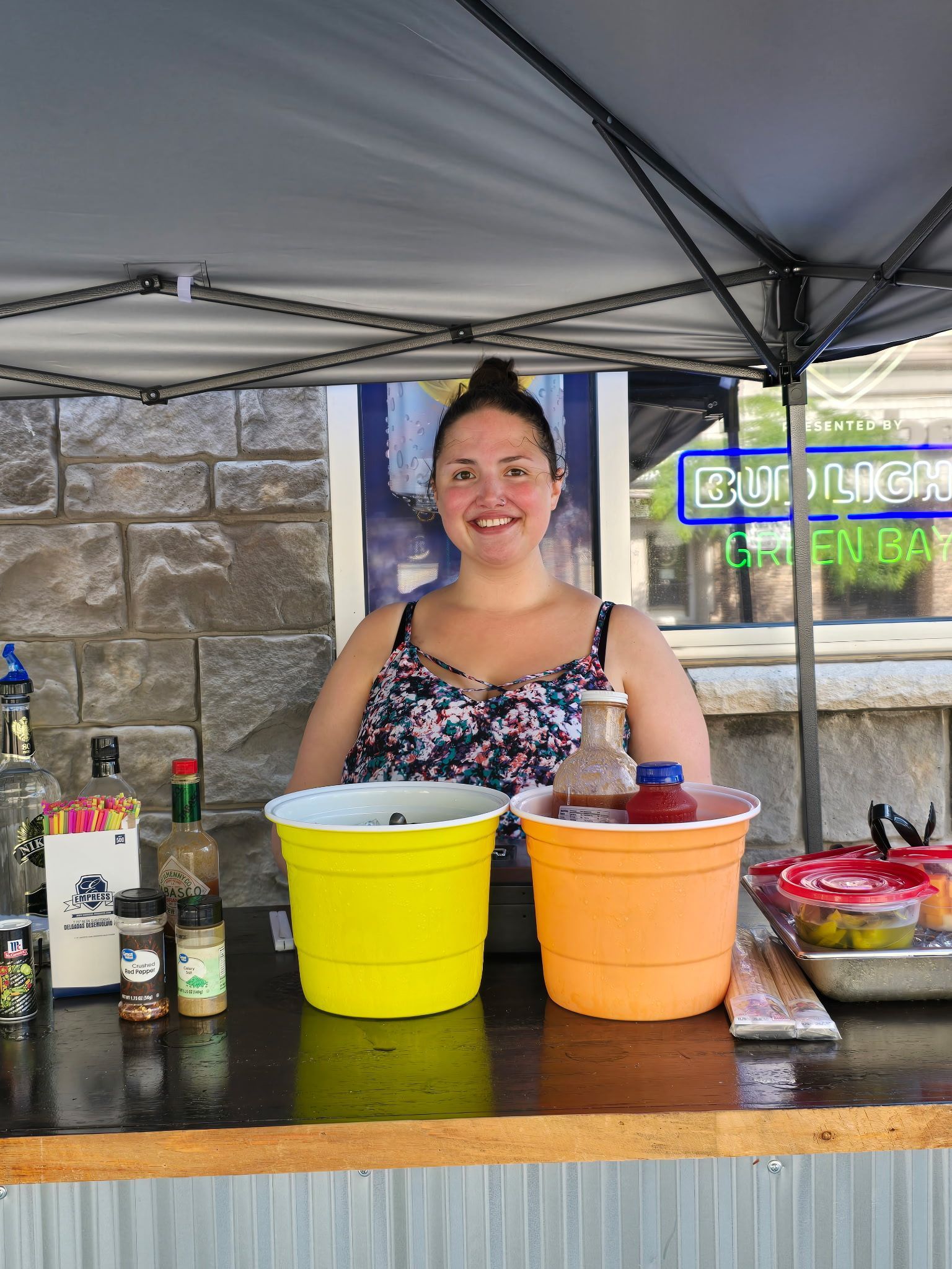 Woman smiling behind a bar, yellow and orange buckets, bottles, and ingredients on counter.