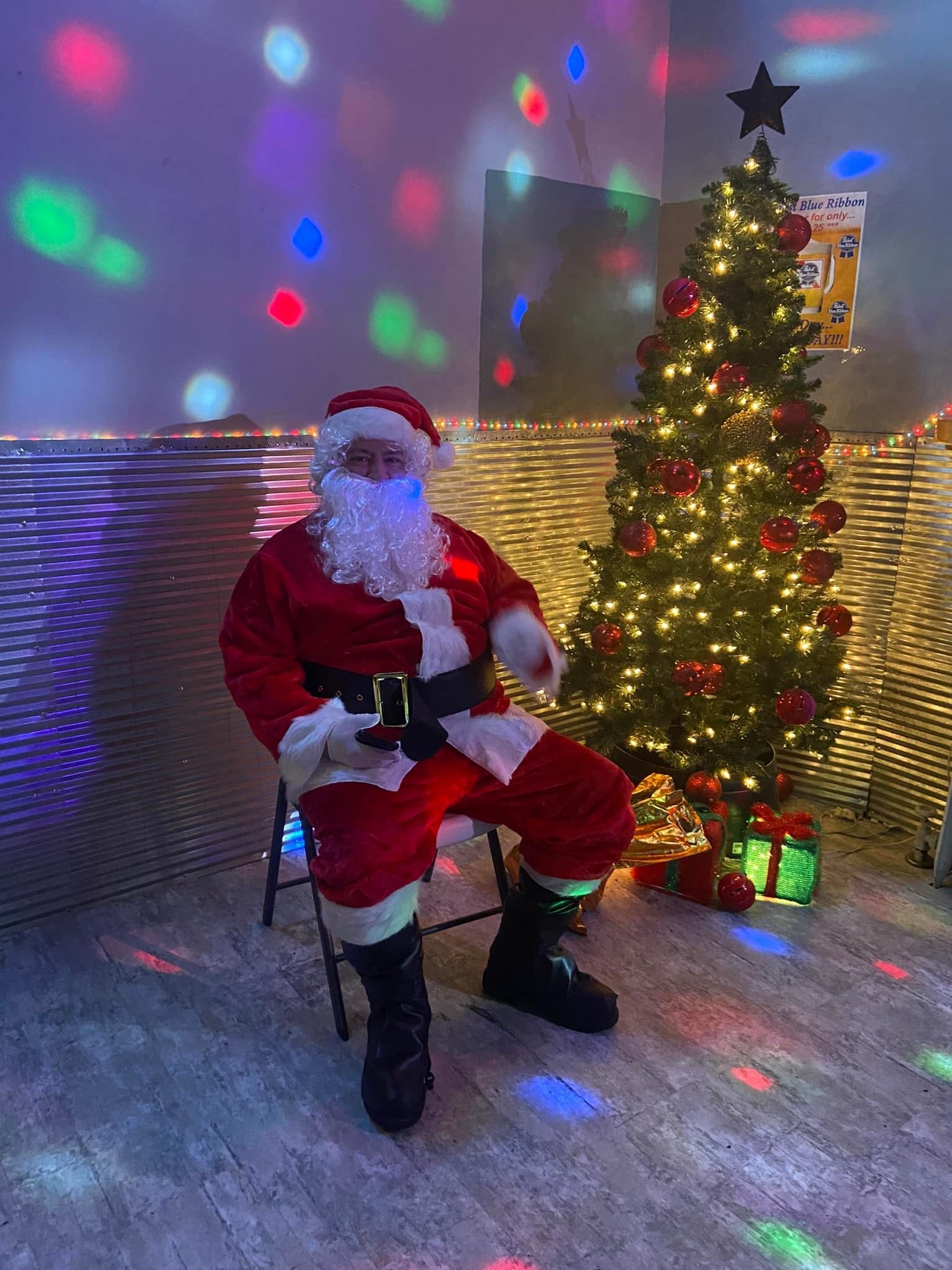 Santa Claus sits on a chair next to a decorated Christmas tree with colorful lights.