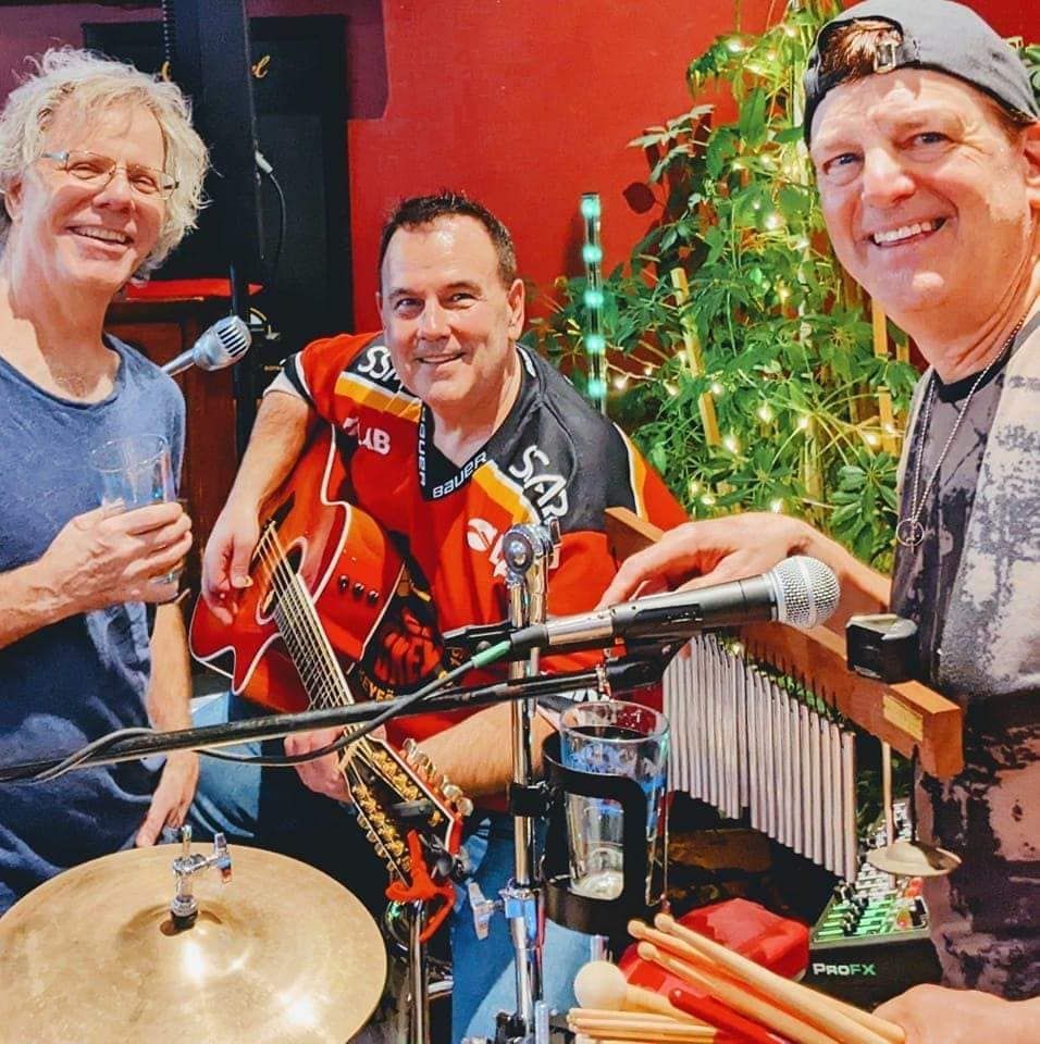 Three musicians pose near instruments and a Christmas tree, smiling.