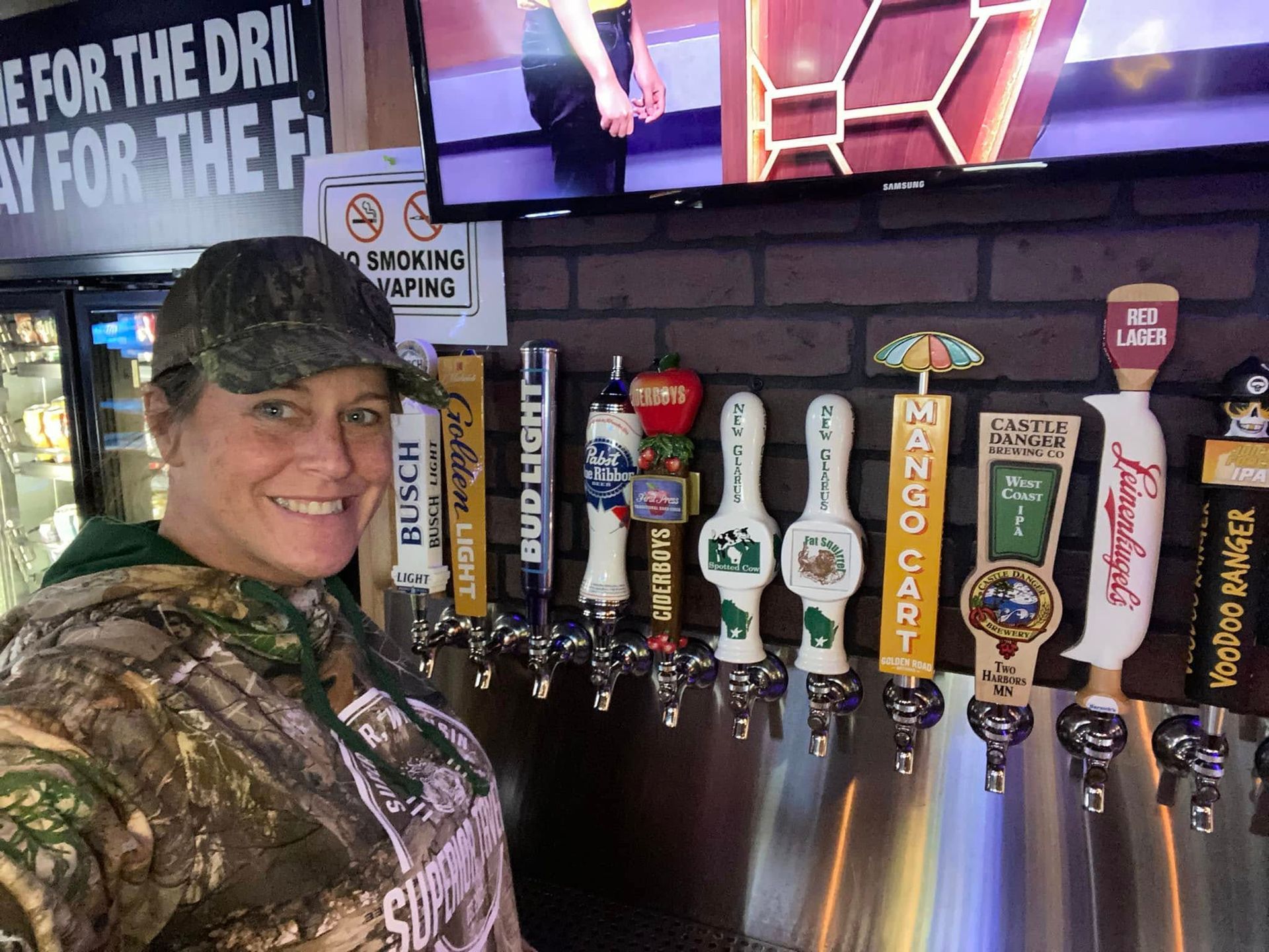 Woman in camouflage at a bar, smiling. Beer taps in foreground, TV on wall.