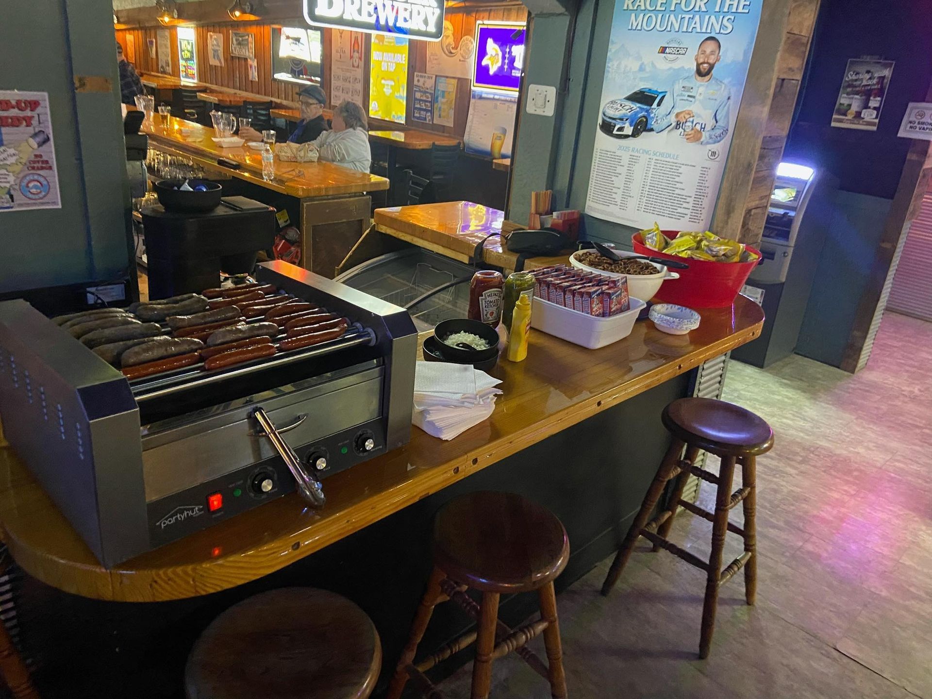 Bar scene: hot dogs on a grill, bar stools, condiments, and a bartender in the background.