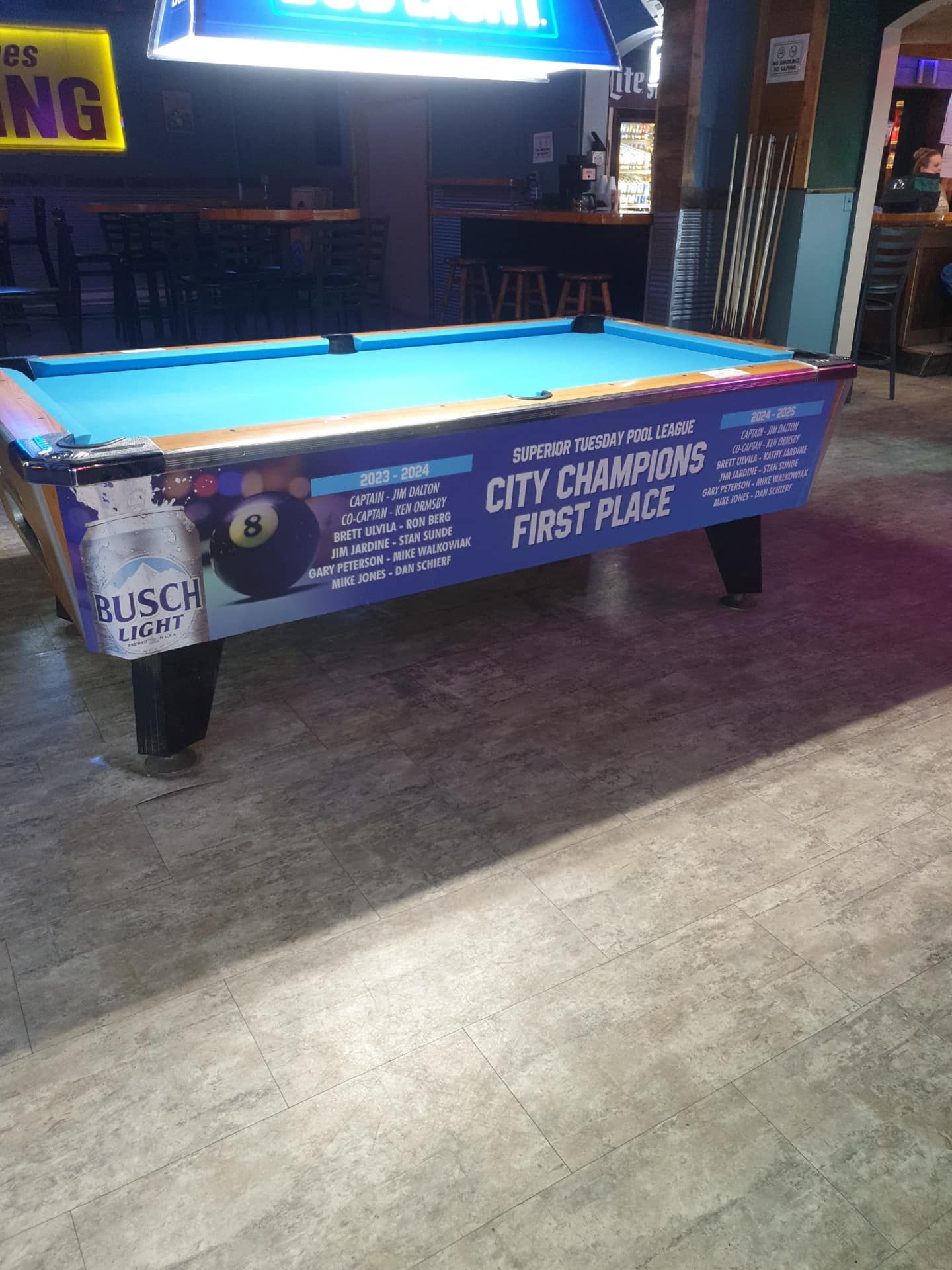 Pool table in a bar with a Busch Light advertisement, blue felt and wood floor.
