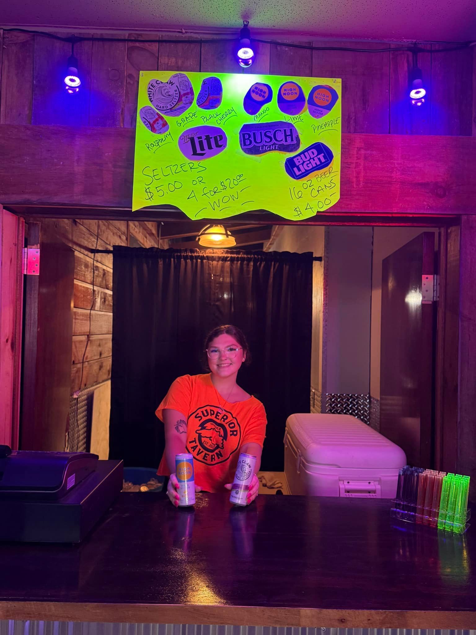 Person at a bar holding two cans, with neon sign and cooler. Lit with purple lights.