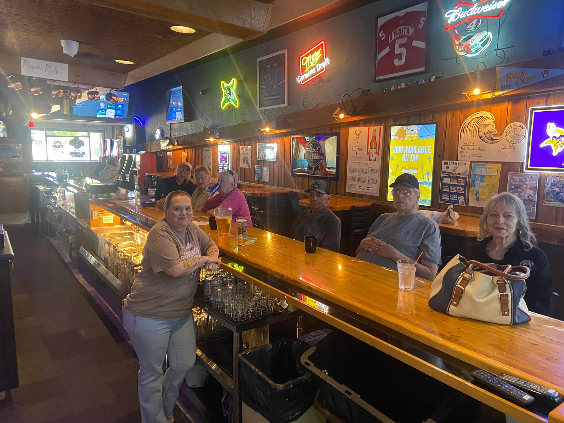 People at a bar, leaning and seated. Wooden bar, drinks, TV screens, neon signs.