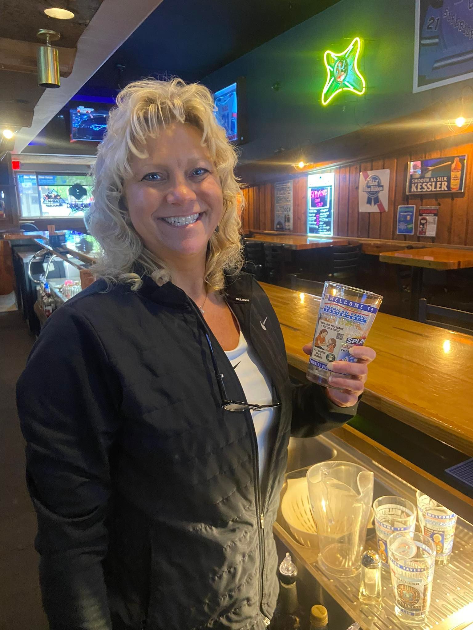 Woman in a bar smiling, holding a decorated glass. Behind her is a bar with glasses and a neon sign.
