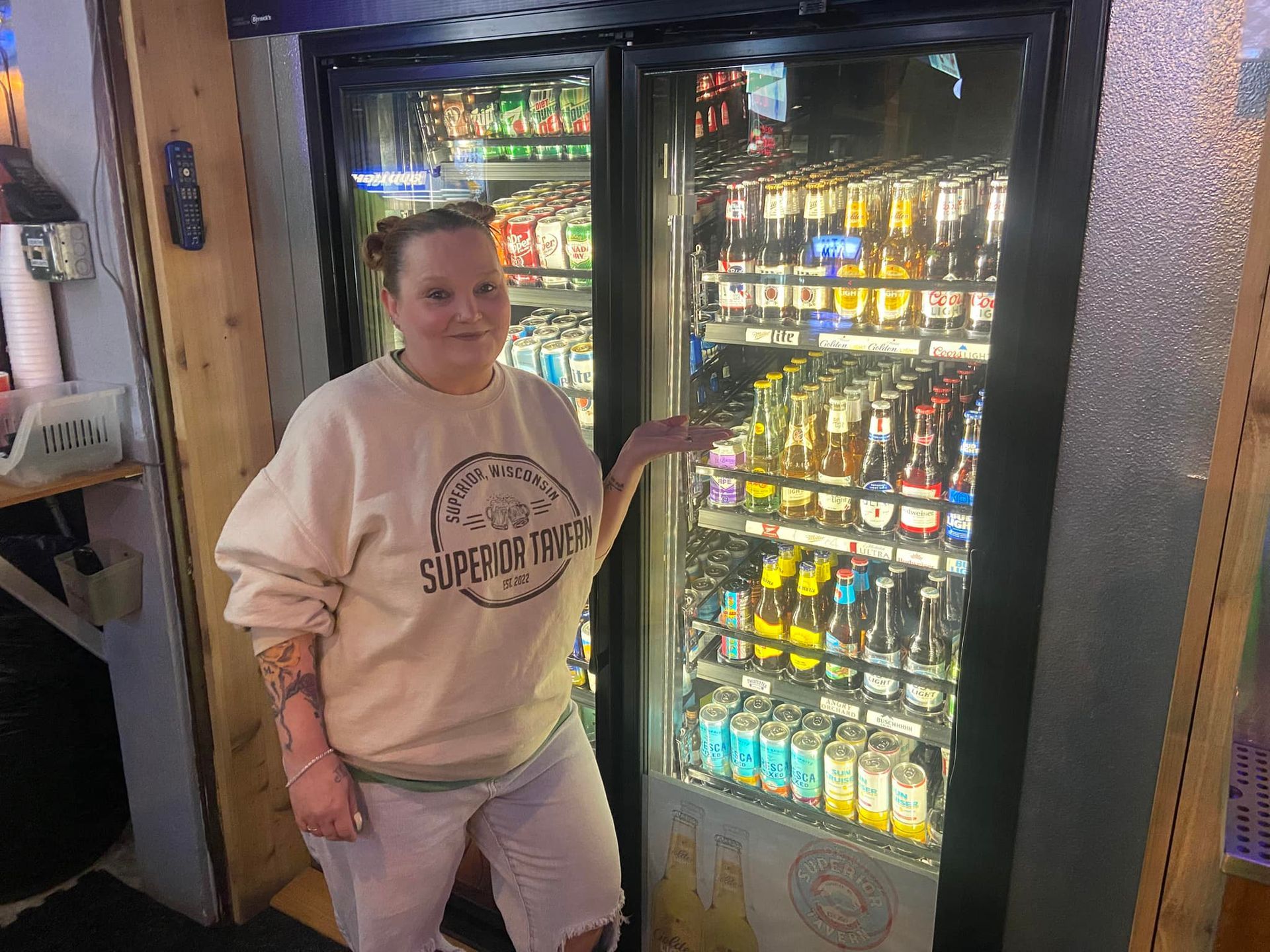 Woman beside a refrigerated beverage display, smiling. Light-colored sweatshirt, tattoos on arm, in a store.