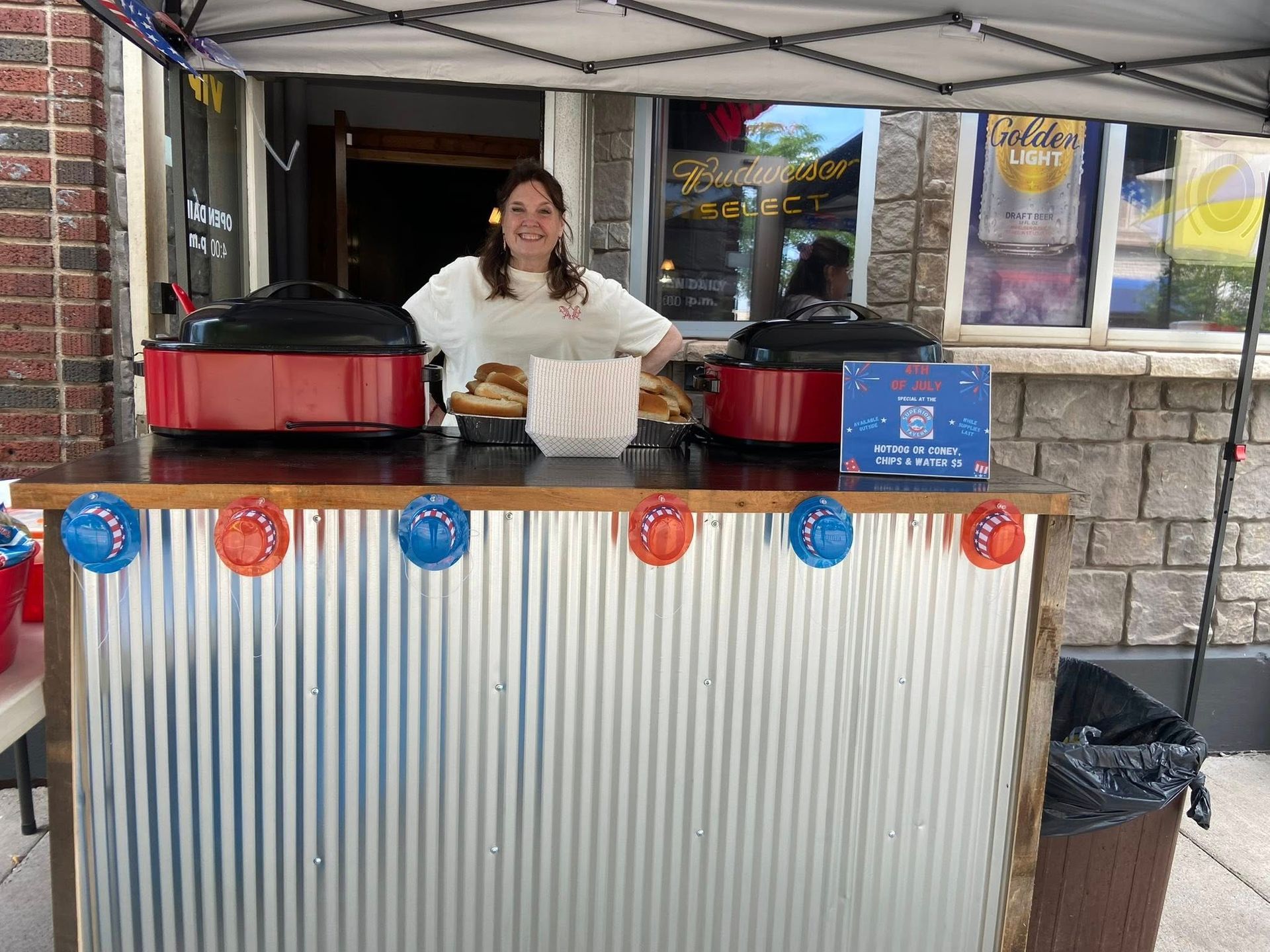 Woman at food stall with red cookers and corrugated metal counter, decorated with red and blue accents.