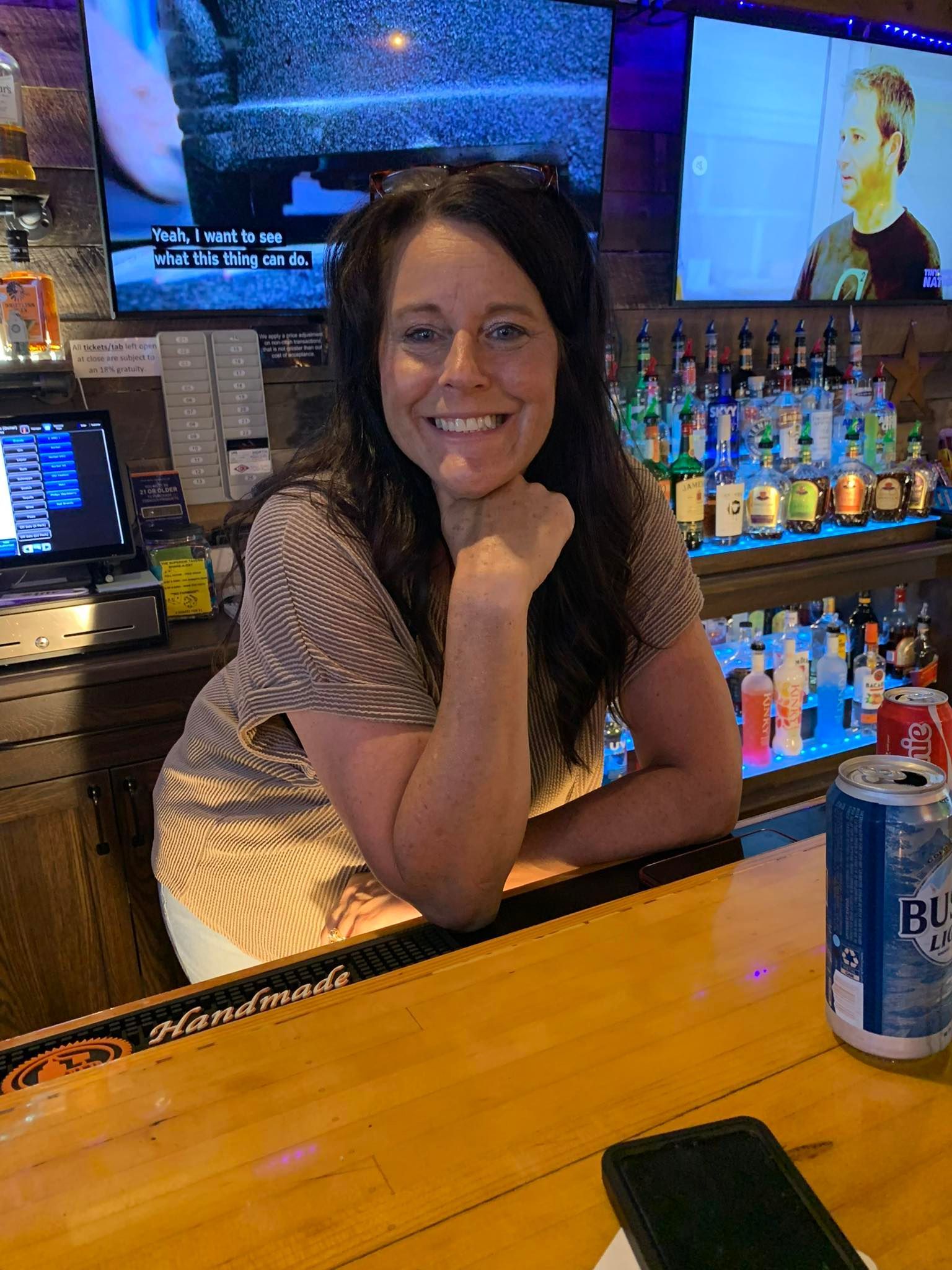 Woman smiling behind a bar, leaning on it. Blue beer can and phone visible. Bottles and TVs in the background.