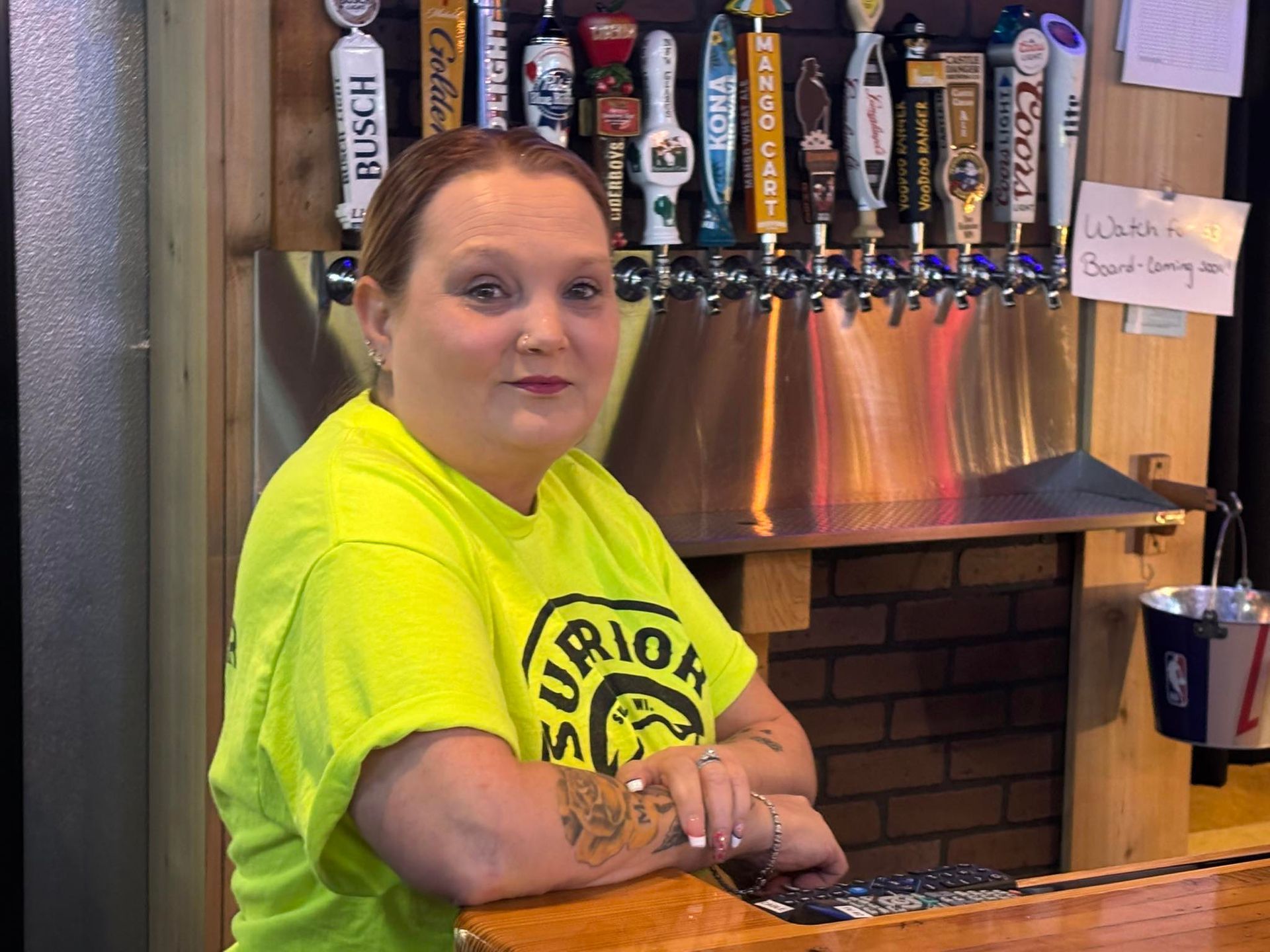 Woman in neon shirt behind a bar, arms resting on counter. Tap handles in the background.