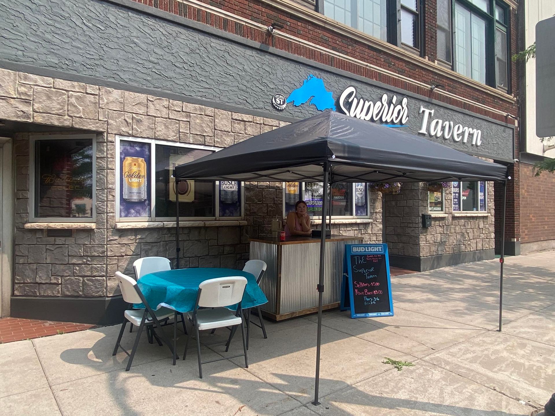 Exterior of Cuderick Tavern with outdoor seating, awning, and a person behind a makeshift bar.