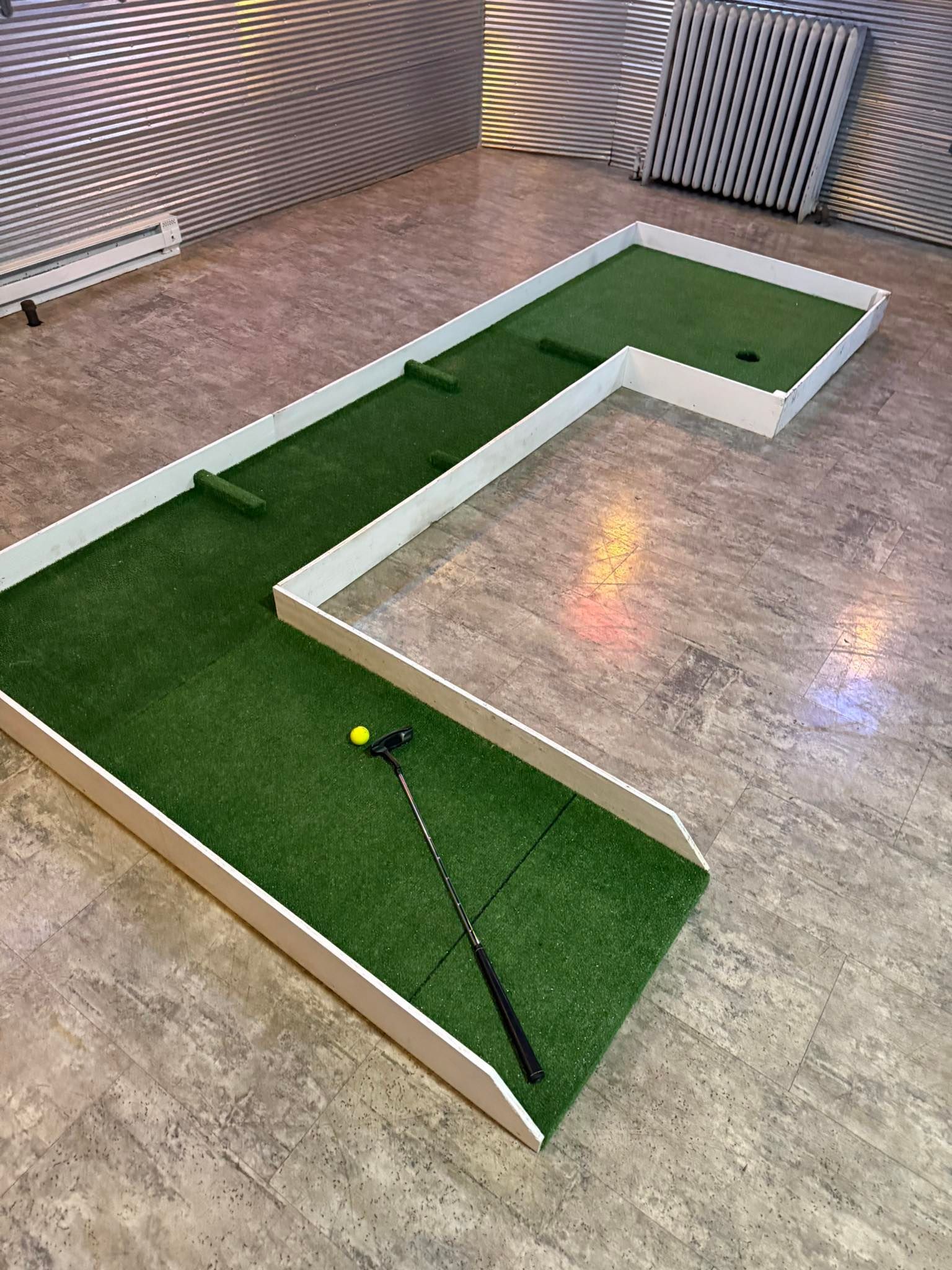 Miniature golf course with green turf, white borders, and a putter with a yellow ball.
