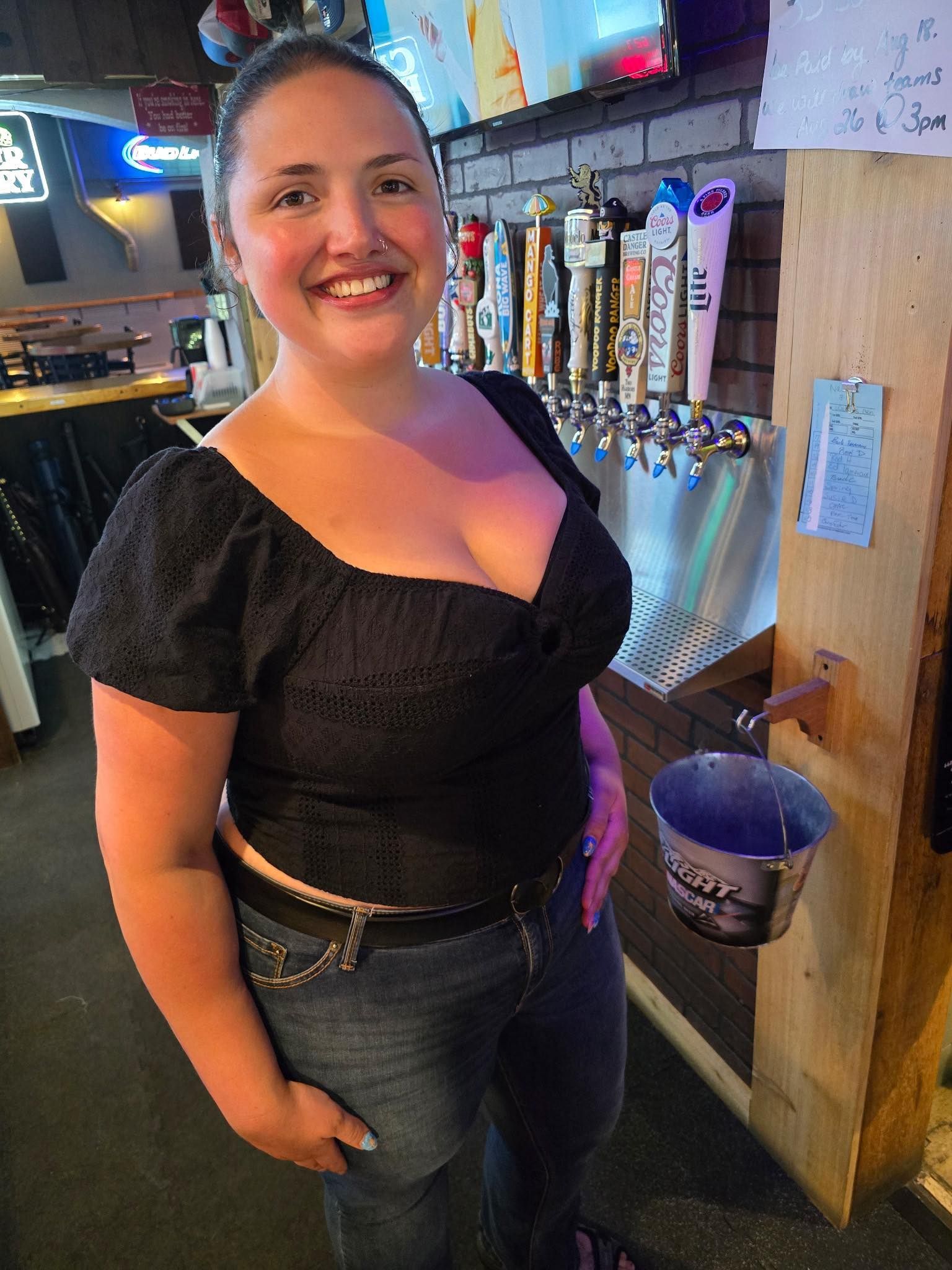 Woman in a black top and jeans smiles in front of a bar, near beer taps and a small bucket.