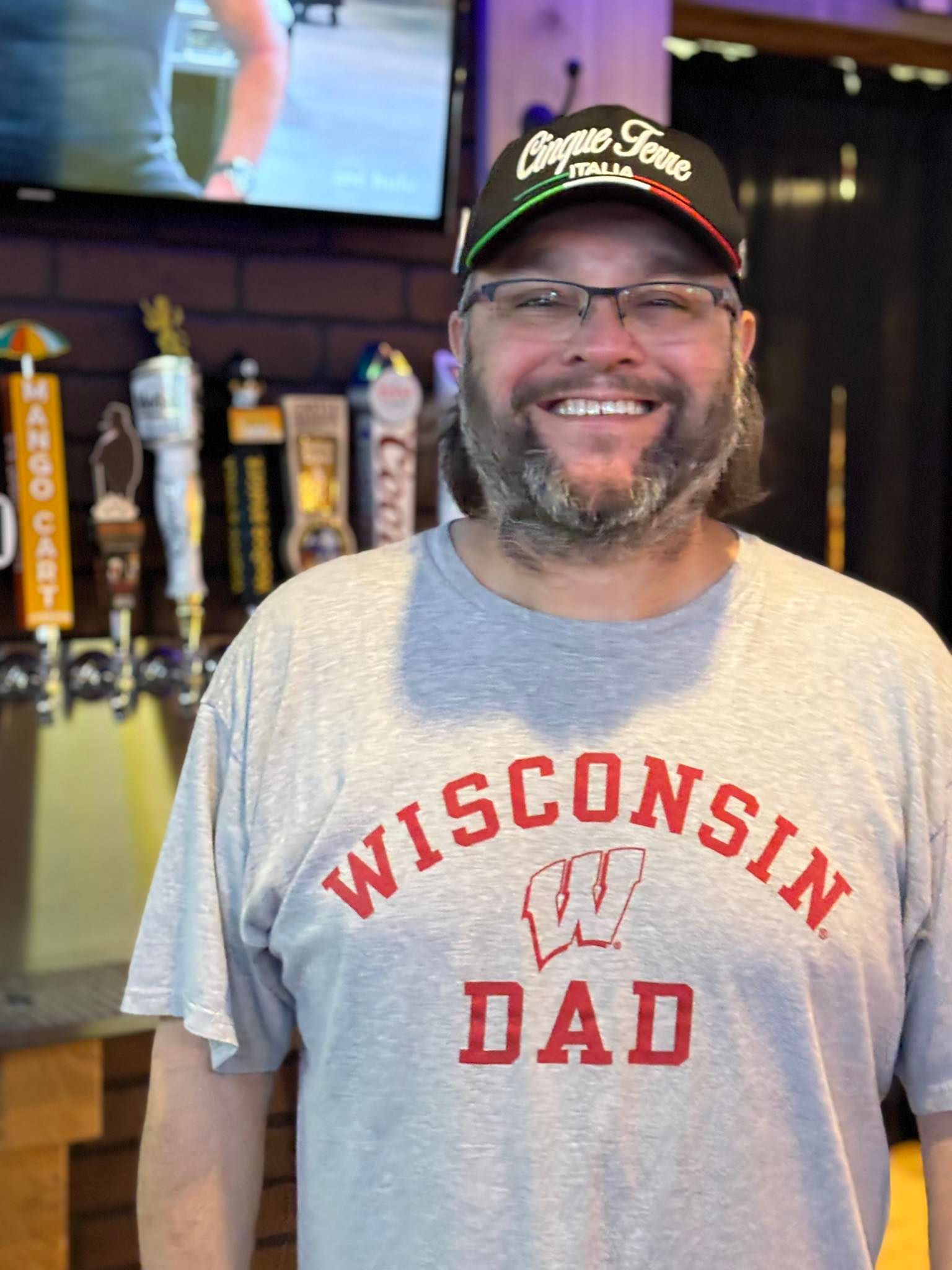 Man wearing a Wisconsin Dad shirt and hat smiles at the camera, standing in front of a bar with taps.