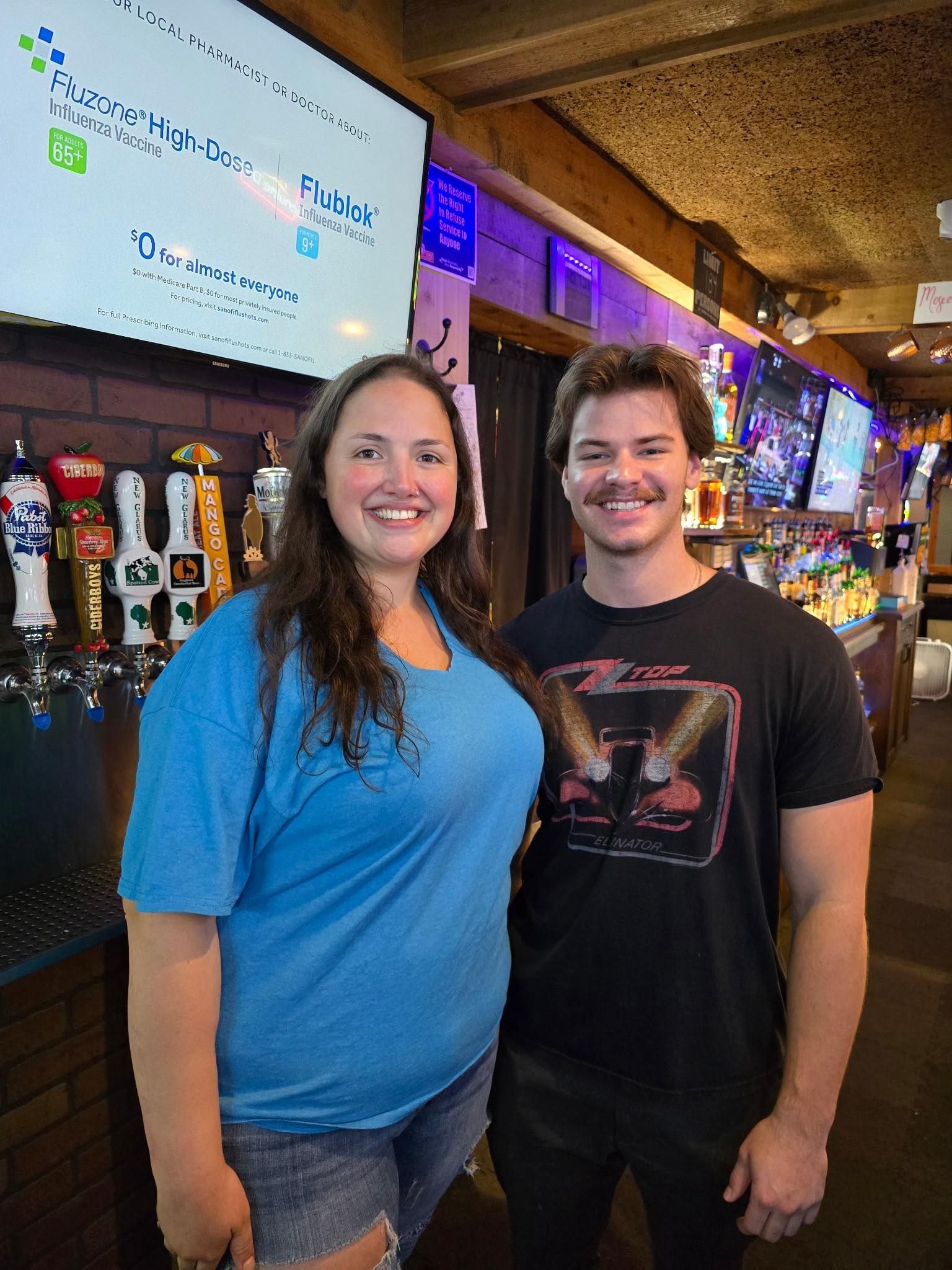 Two people smiling, standing at a bar. Woman in blue shirt and shorts, man in black shirt. Dark interior, taps behind.