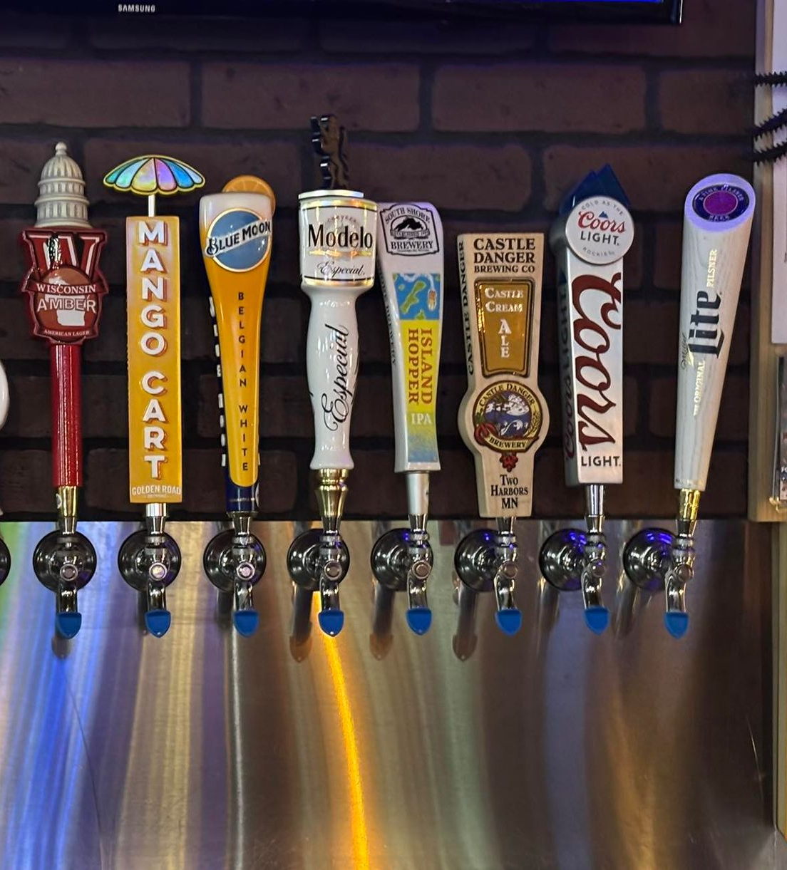 Row of beer taps with various logos against a brick wall background.
