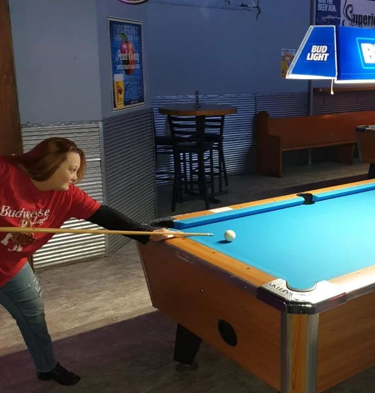 Woman in red shirt playing pool in a bar. She is aiming with a pool cue at the white ball.