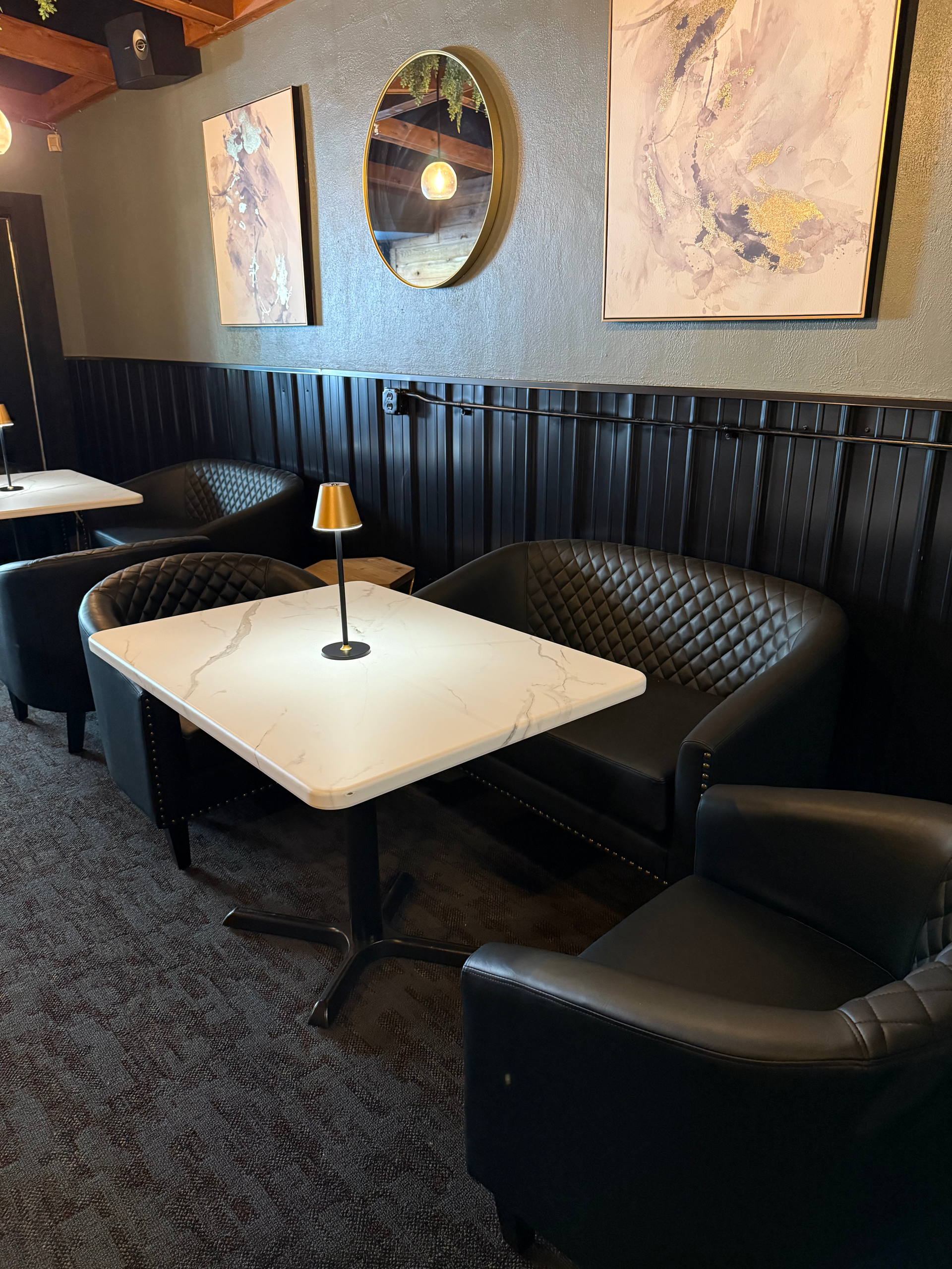 A dimly lit restaurant seating area with black booths, chairs, and tables. Decorative wall art and a mirror are present.