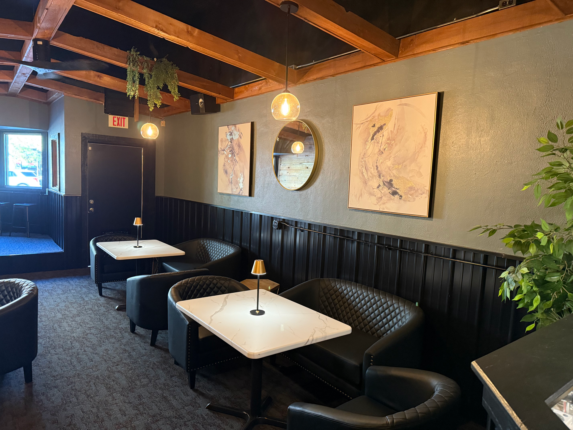 Interior of a dimly lit bar with black leather seating, tables, and artwork on gray walls, with hanging lights.