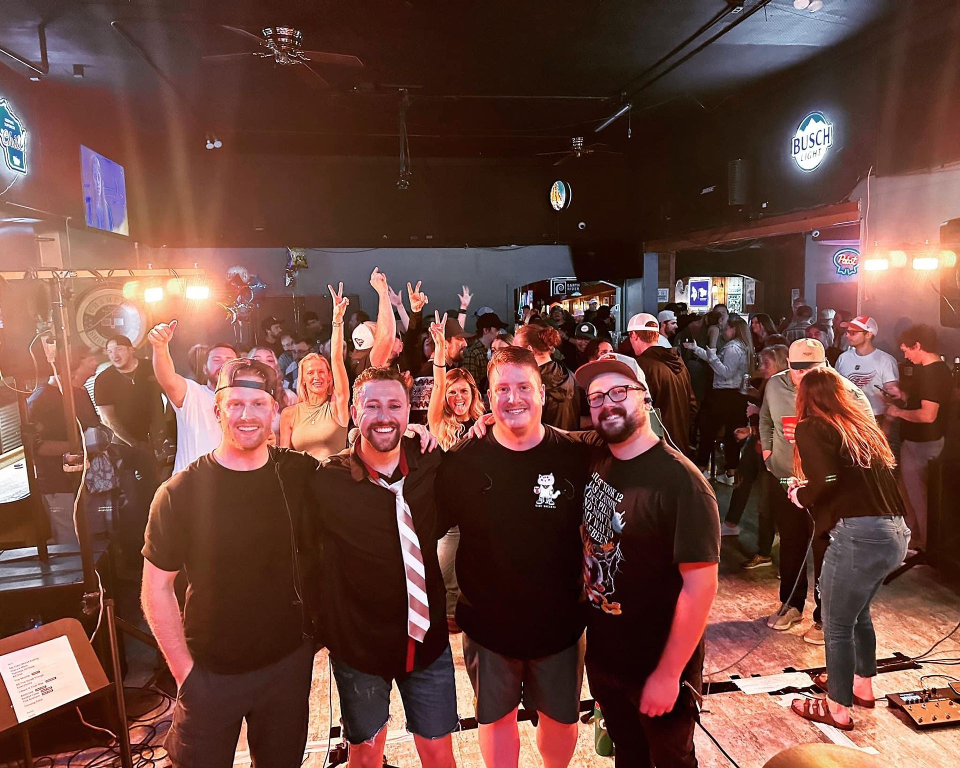 Band onstage with crowd in a dimly lit venue. Four men in front, people in background cheering.