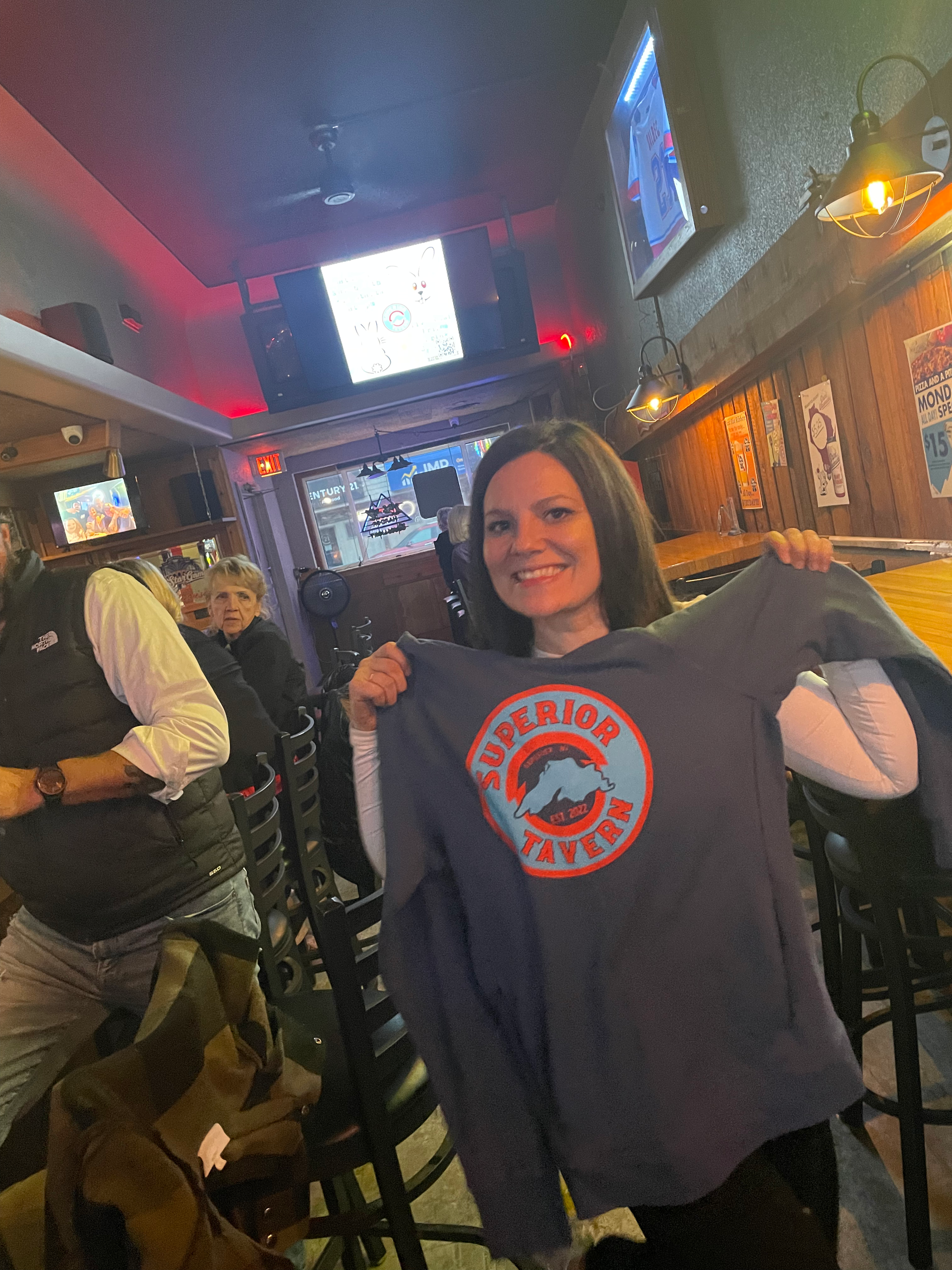 Woman holding up a navy sweatshirt in a bar; logo reads 
