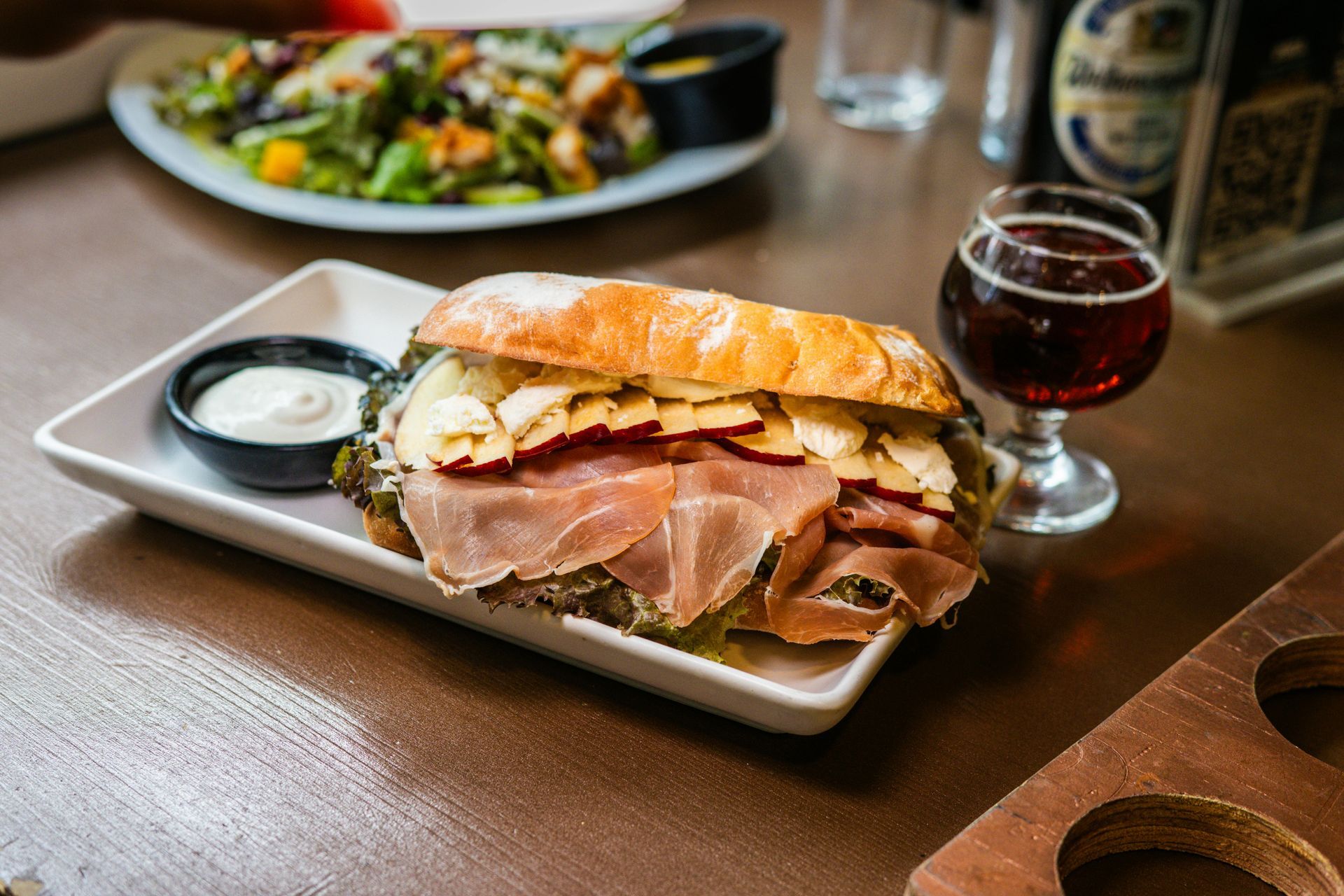 Sandwich with prosciutto and cheese, side of sauce, salad, and a glass of dark beer on a table.
