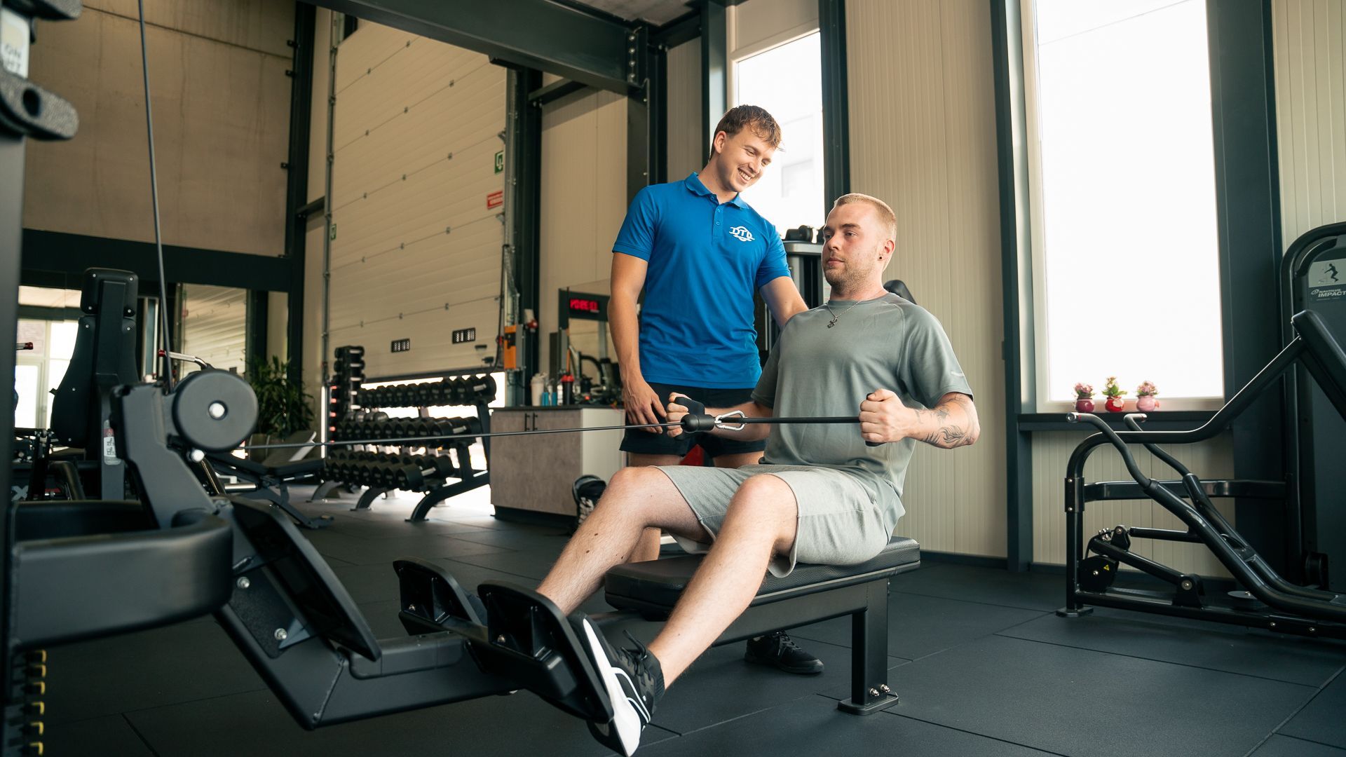 Een man zit op een roeimachine in een sportschool, terwijl een man achter hem staat.