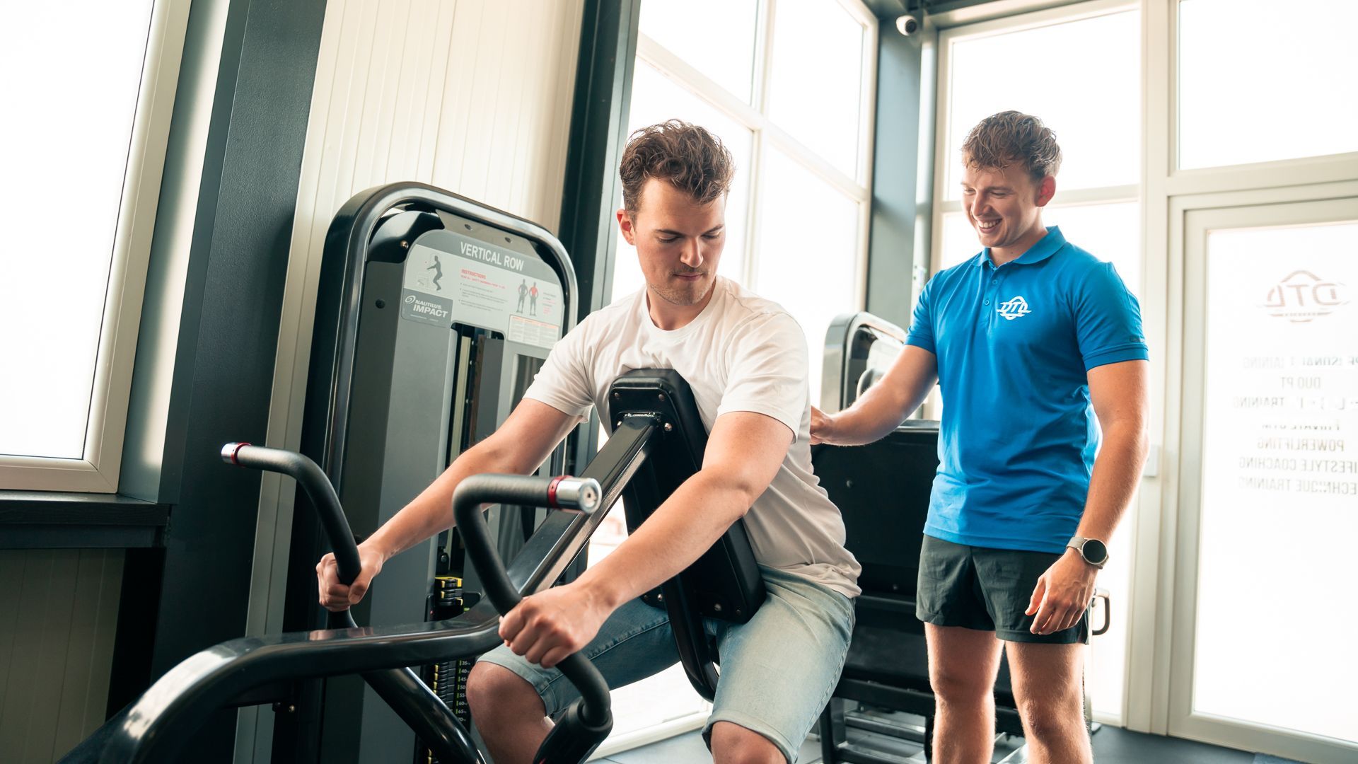 Een man fietst in een sportschool op een hometrainer, terwijl een andere man toekijkt.