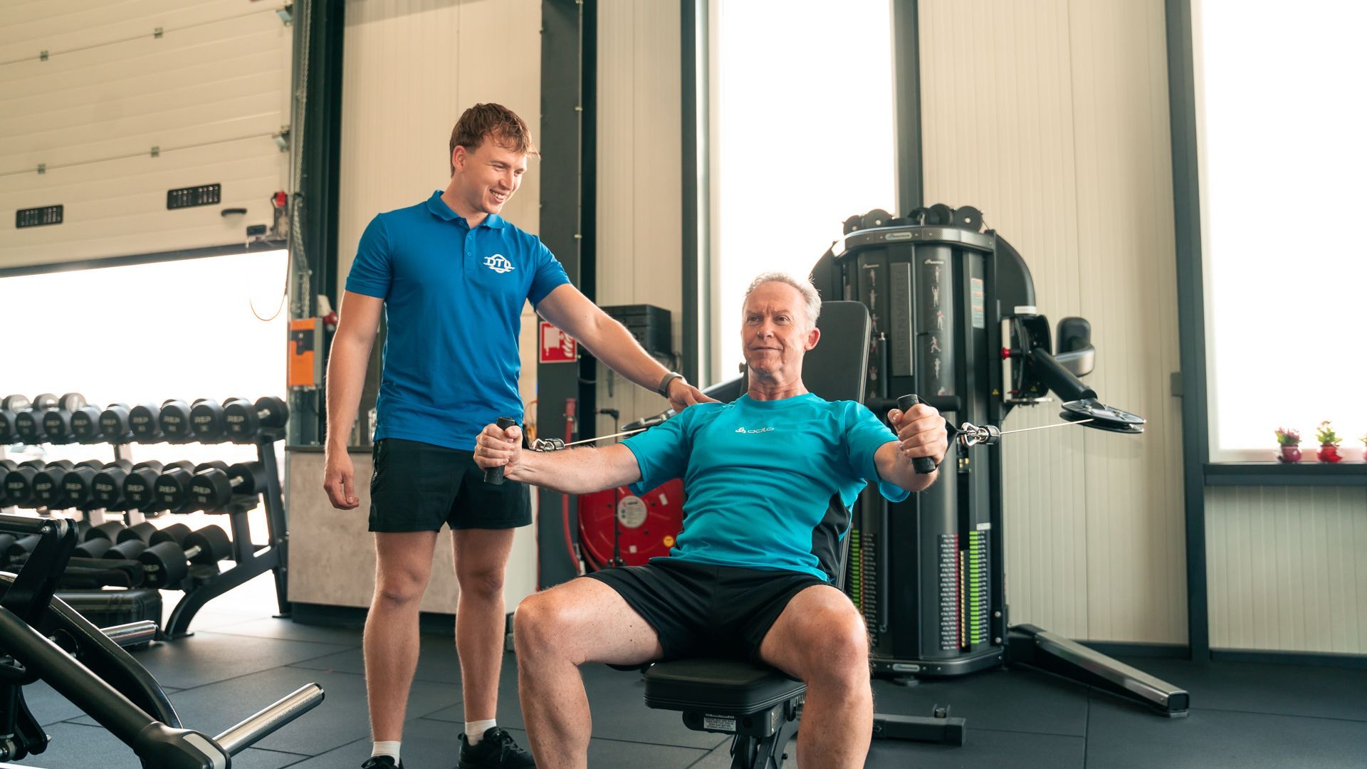 Een man staat naast een man die op een bankje zit in een sportschool.