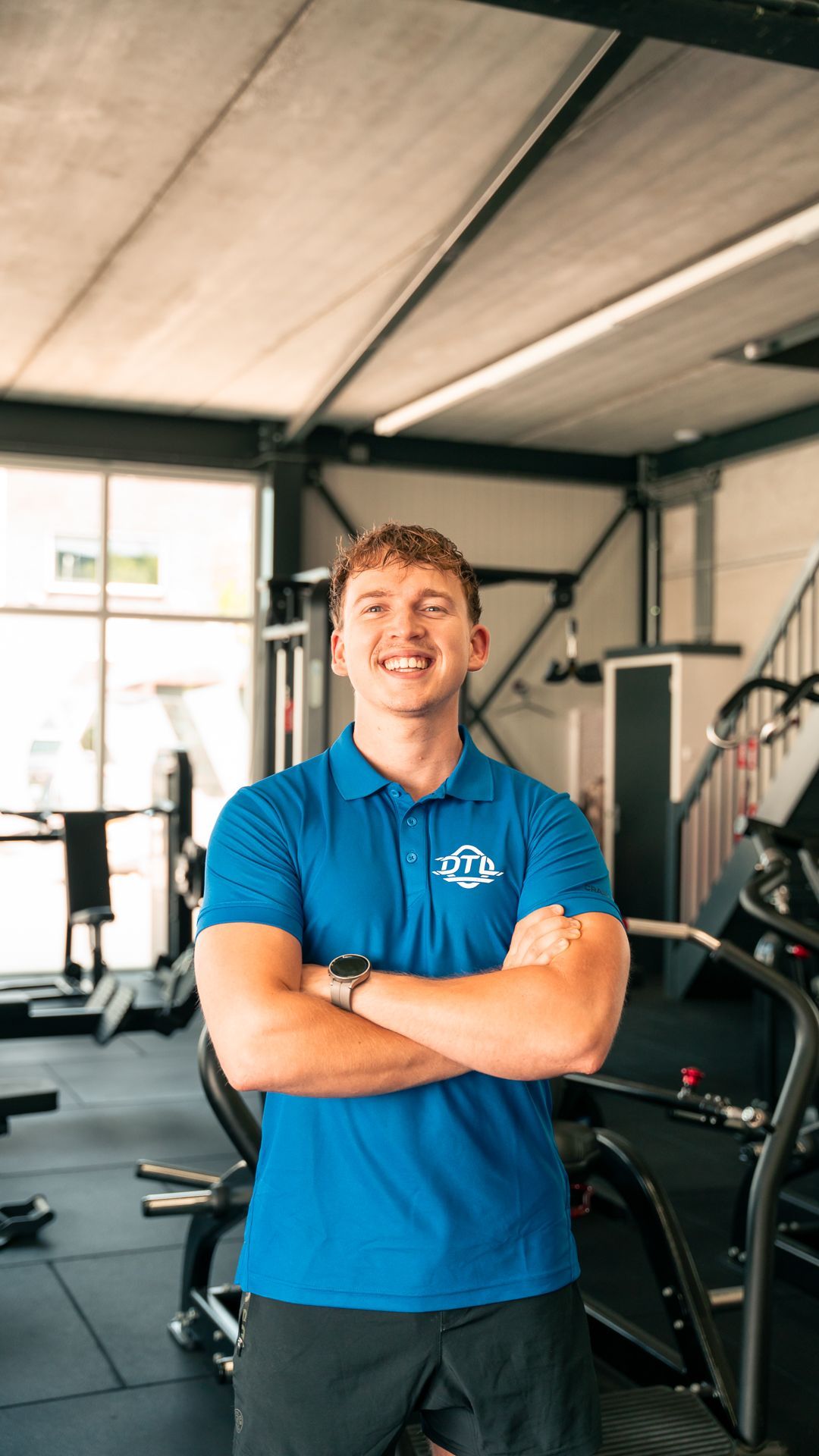 Een man in een blauw shirt staat in een sportschool met zijn armen over elkaar.