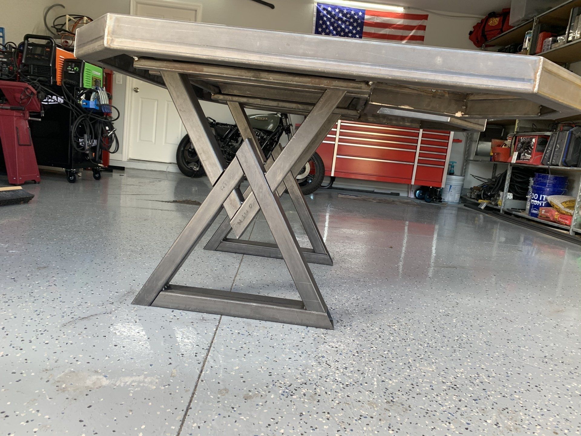 Steel-framed table with X-shaped legs in a garage, with an American flag in the background.