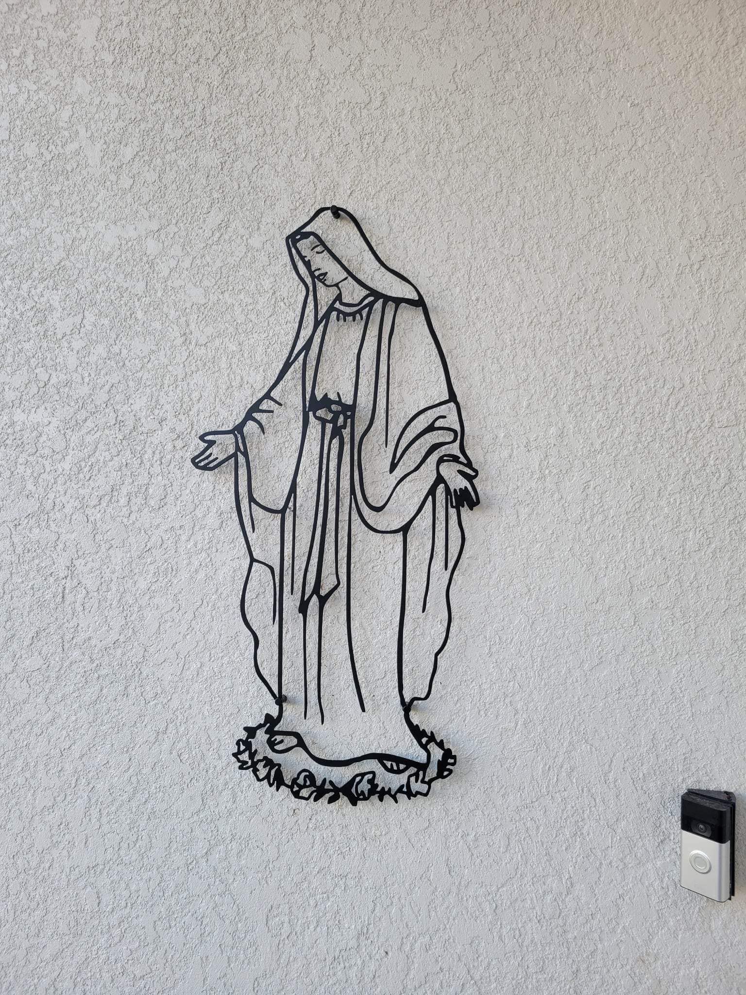 Metal outline of Virgin Mary on textured white wall.