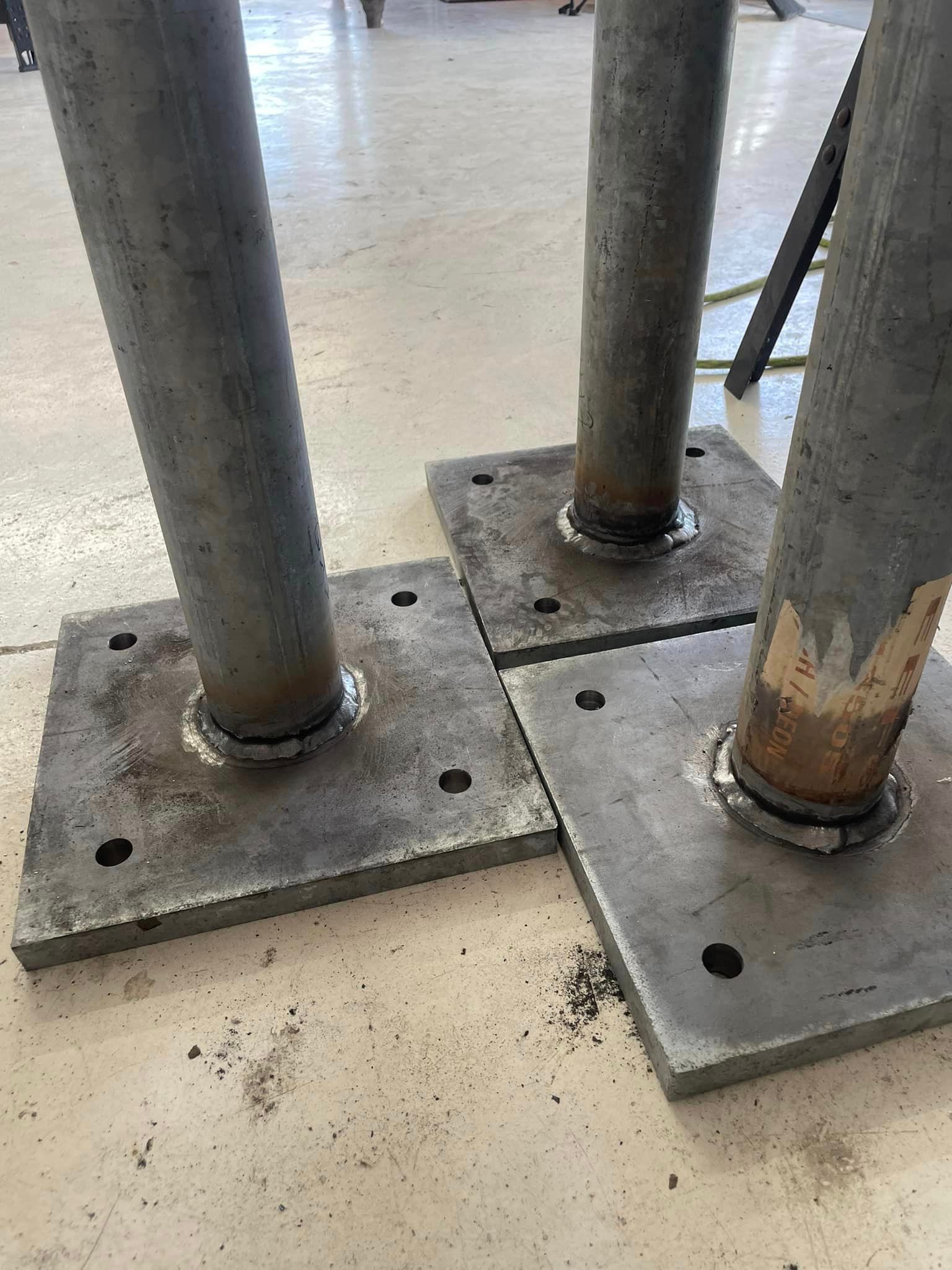 Three steel poles welded to square base plates with bolt holes, on a concrete floor.