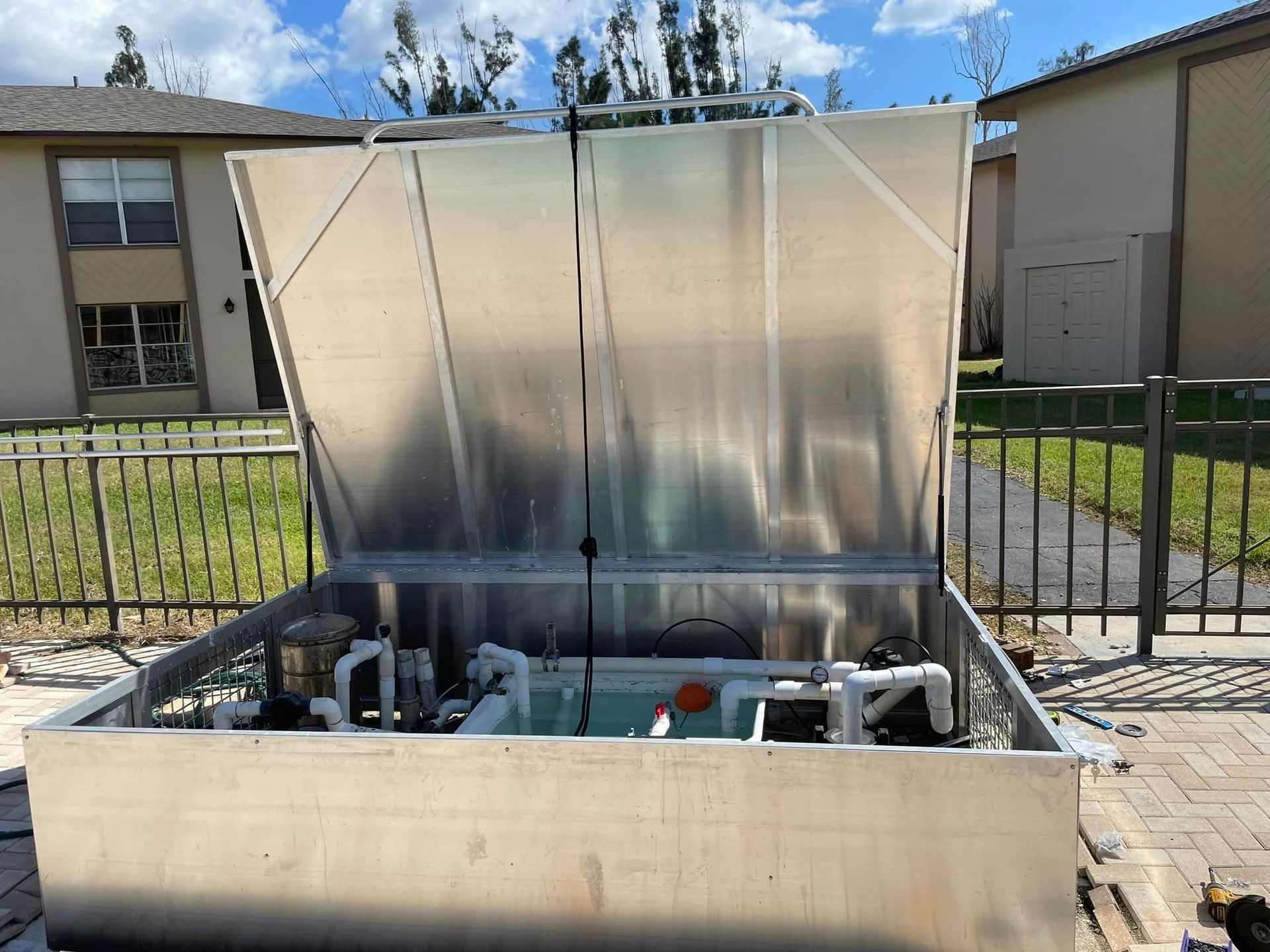 Silver pool equipment enclosure with open lid, revealing pumps and plumbing. Outdoors, near a fence and buildings.
