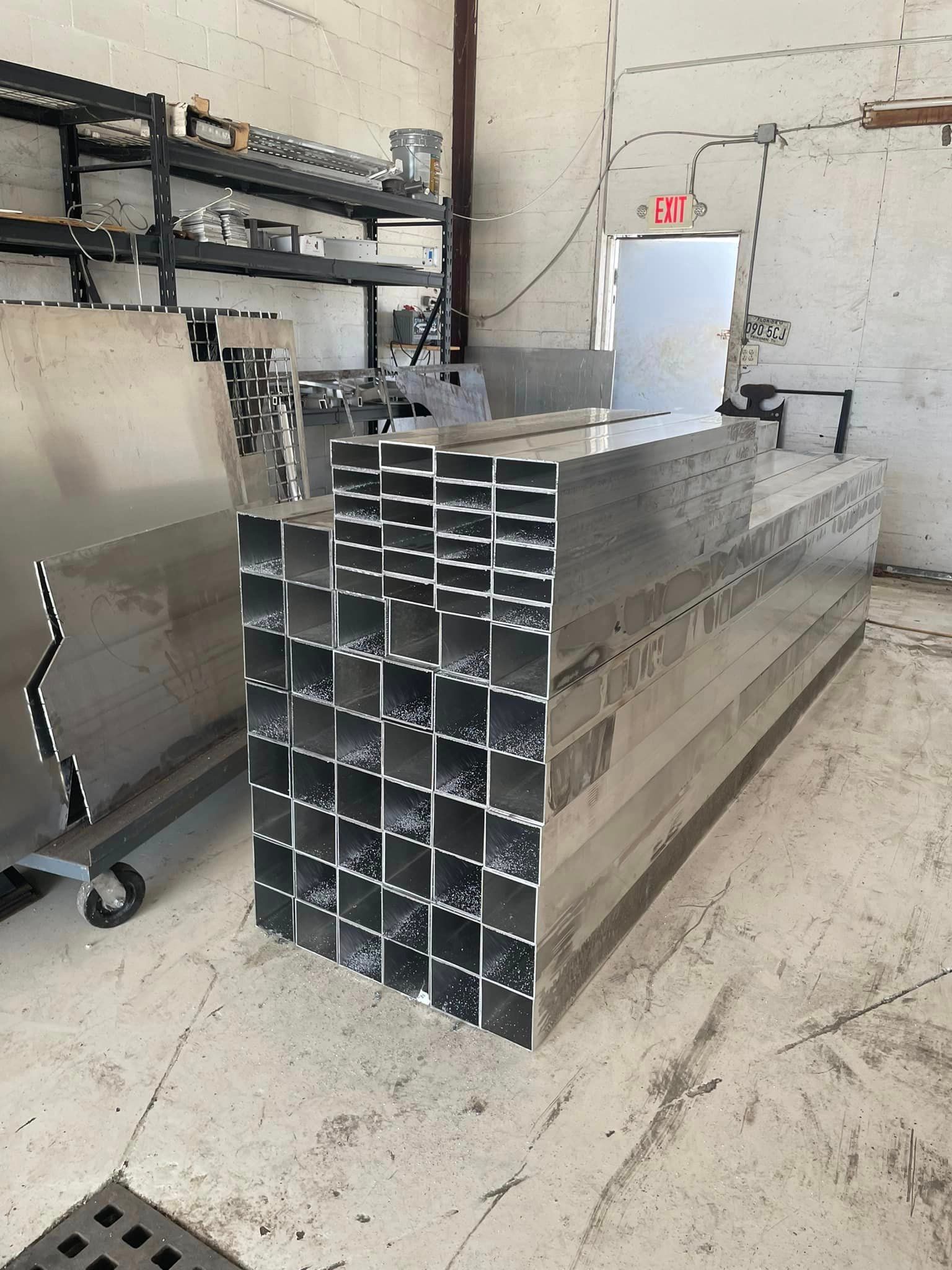 Aluminum square tubing stacks inside a workshop; a metal cart and shelving are visible.