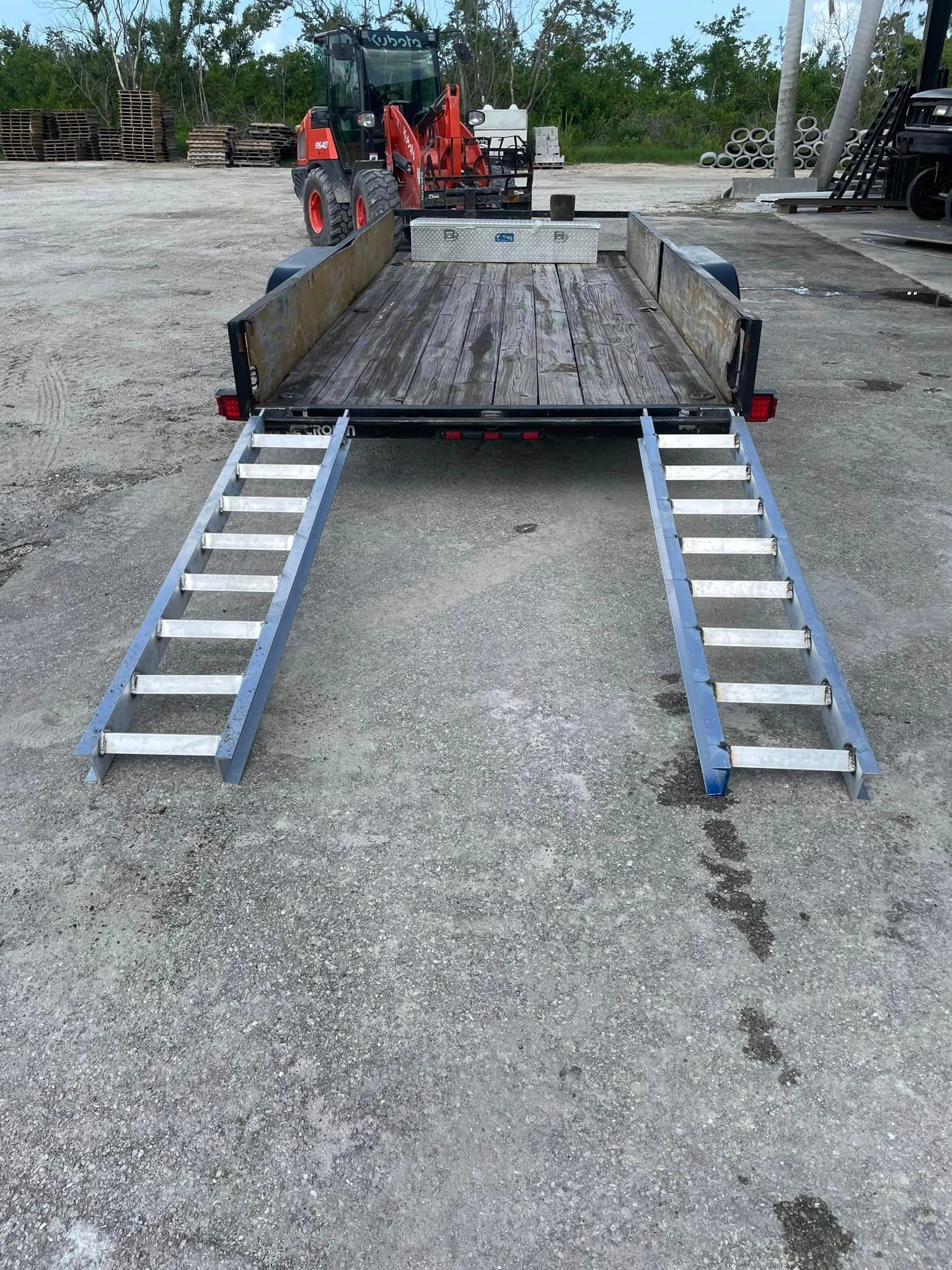 Trailer with loading ramps, a tractor in the background.  Gray concrete, sunny day.