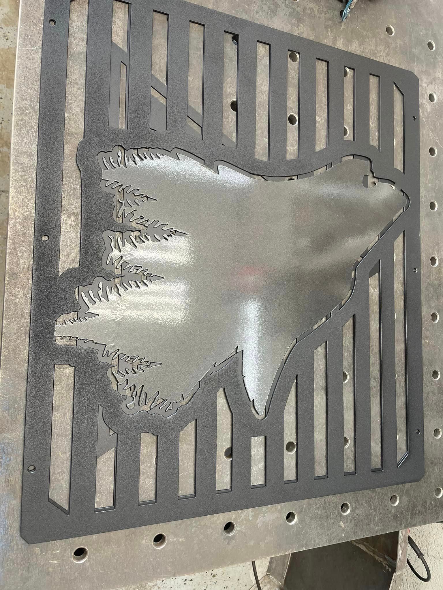 Metal cut-out with bear shape, trees, and vertical bars; dark gray on a metal table.
