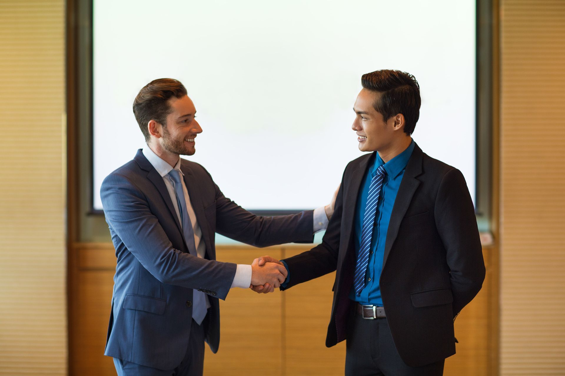 Two men in suits and ties are shaking hands in front of a projection screen.