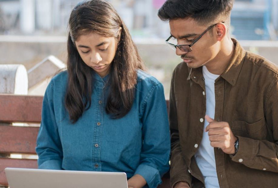 A man and a woman are sitting on a bench looking at a laptop.