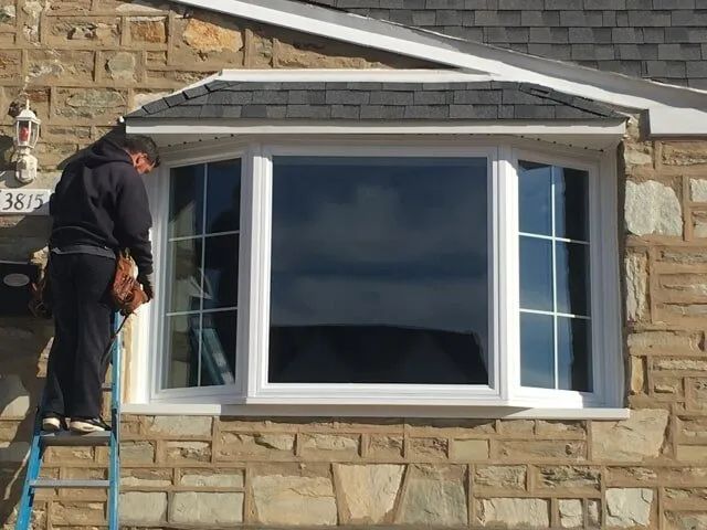 A man is standing on a ladder fixing a window on a brick wall.