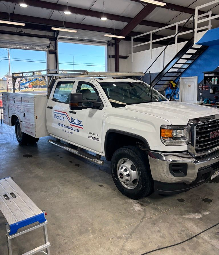 A white GMC work truck with a utility bed and roof rack parked inside an industrial shop.