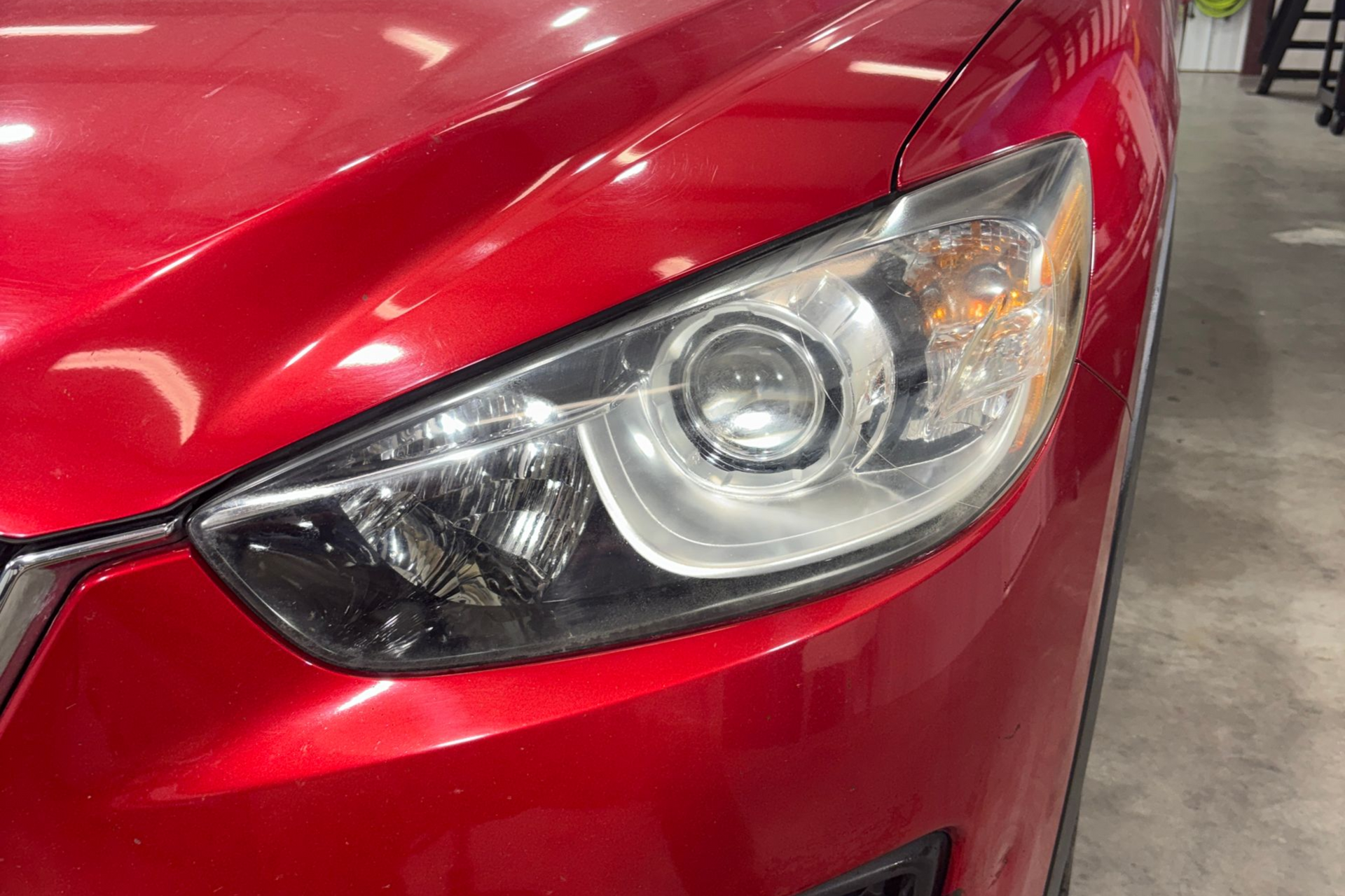 Close-up of a red Mazda CX-5 SUV's front passenger side, showing the headlight, fog light, and hood in a garage.
