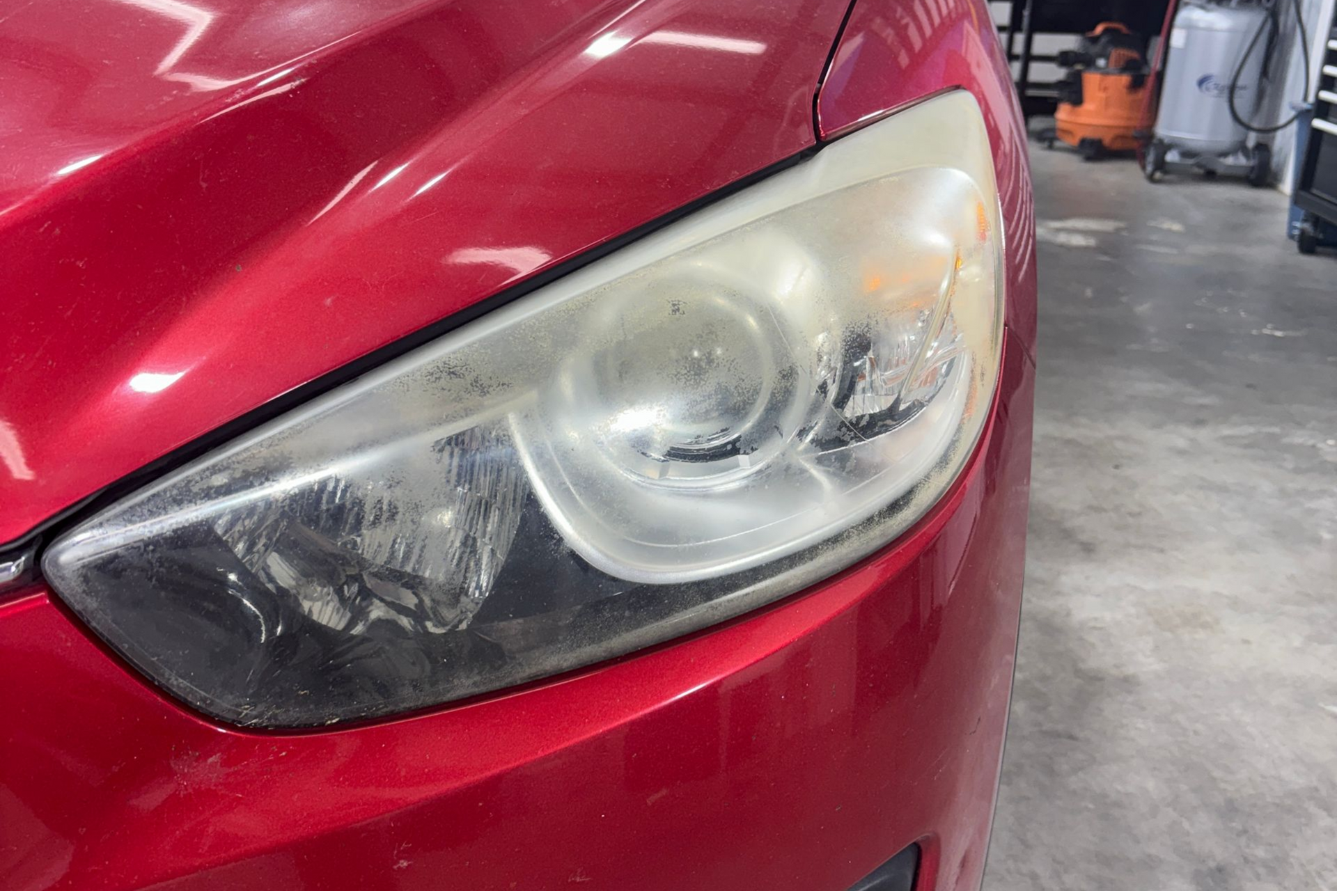 Close-up of a red car’s headlight assembly showing signs of oxidation and yellowing in an auto shop garage.